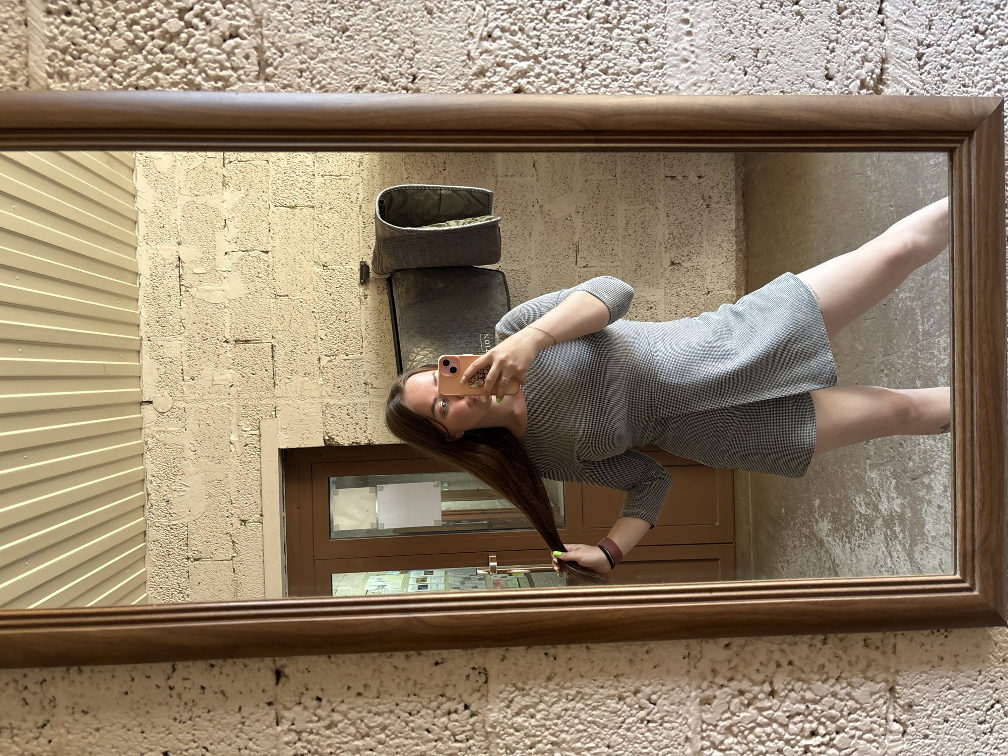 A woman is taking a selfie in a rectangular mirror with a wooden frame. The reflection shows her wearing a grey dress, holding a pink smartphone. Her long brown hair extends down to her wrist. The background reflected in the mirror shows a textured wall, a grey padded object, and a brown door with a window. The location is not discernible from the image.