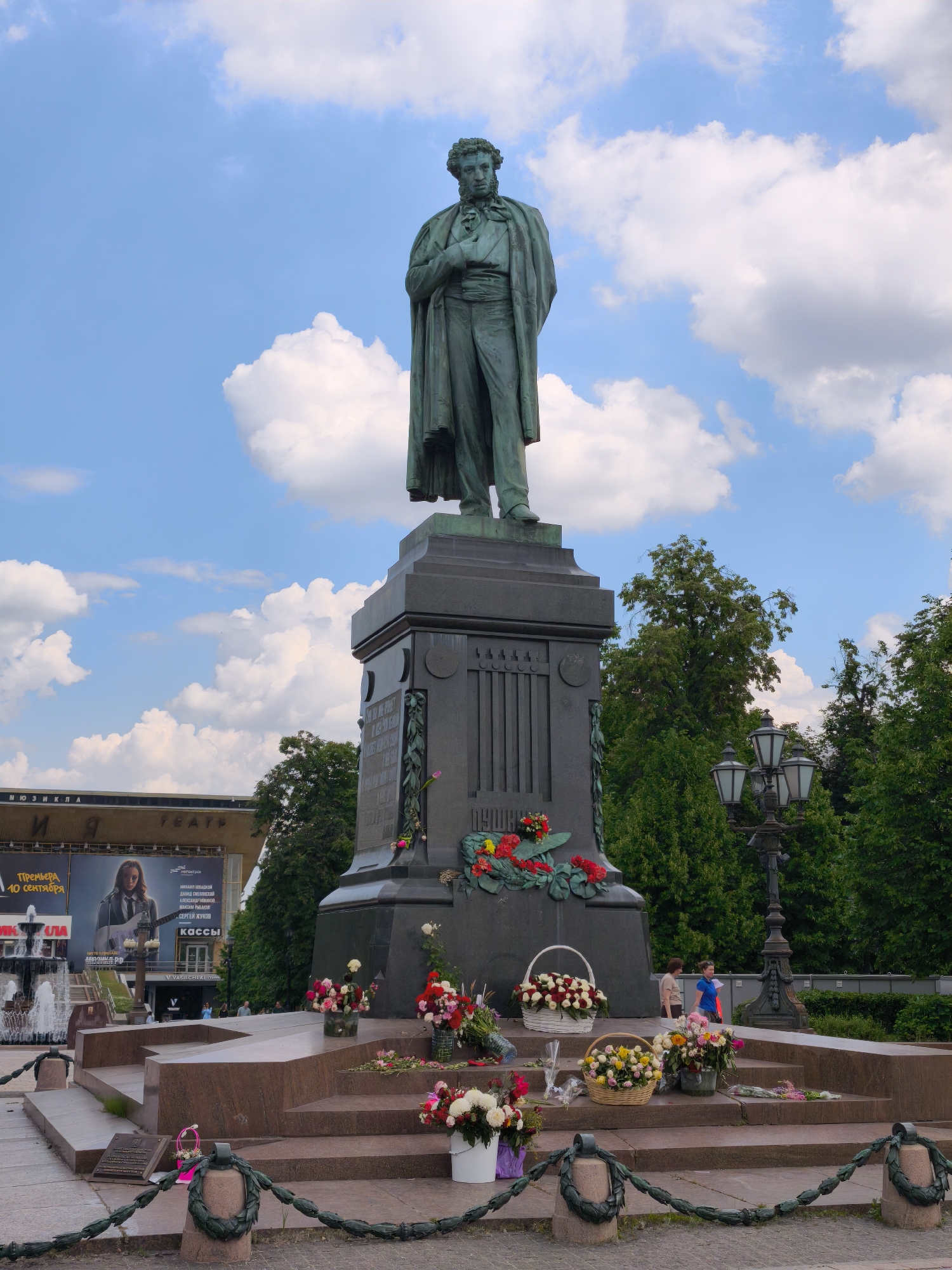 The photograph captures a bronze statue of Alexander Pushkin, a prominent Russian poet, located in Pushkinskaya Square, Moscow, Russia. The statue stands on a granite pedestal, adorned with floral wreaths and bouquets at its base, suggesting a recent commemoration or memorial event.
In the background, the facade of a theater building is visible, featuring a large advertisement poster. The sky is partly cloudy, with a mix of blue sky and white clouds, illuminated by daylight. Several trees are present, framing the scene and adding a natural element to the urban environment.
In the middle ground, two women are walking near the base of the statue, one wearing a light-colored top and skirt, and the other a blue shirt and dark pants. A vintage street lamp with multiple lights stands to the right of the statue. The foreground shows the steps leading up to the monument, with more floral arrangements, including baskets of flowers and individual stems, placed on them. A decorative chain with green garland-like elements cordons off the area around the monument. A fountain is visible to the left of the frame.