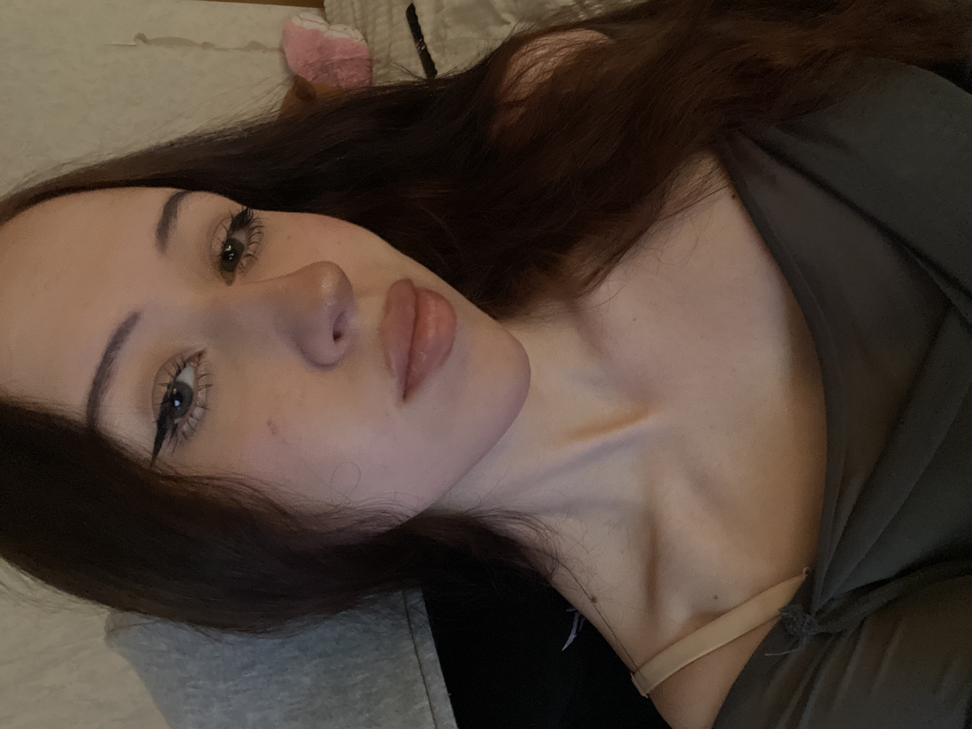 The image displays a head-and-shoulders portrait of a young female in an indoor setting. The specified location is the city of Votkinsk, Russia, although no specific visual cues within the image confirm this geographical detail. The subject has long, dark brown hair, parted centrally, and light-colored eyes (appearing hazel or green). She wears winged black eyeliner and mascara, and her lips are full, appearing to have a light gloss or lipstick. A small dark mole or freckle is visible below her right eye, and another small mole is present on her upper left chest near the collarbone. She is wearing a dark, possibly olive-gray, off-the-shoulder top or dress, with a beige bra strap visible over her left shoulder. The subject's expression is neutral as she looks directly at the camera.
The background on the left side of the image features a textured gray fabric, possibly a blanket or hoodie, and a darker, undefined surface below it. On the right side, a light-colored, textured wall (possibly wallpaper) is visible, along with a portion of a white quilted surface, likely a bed or couch. A pink, soft object, possibly a pillow or stuffed animal, is partially visible on the white surface, along with a slender dark object. The lighting is dim and soft, suggesting an artificial indoor light source.
