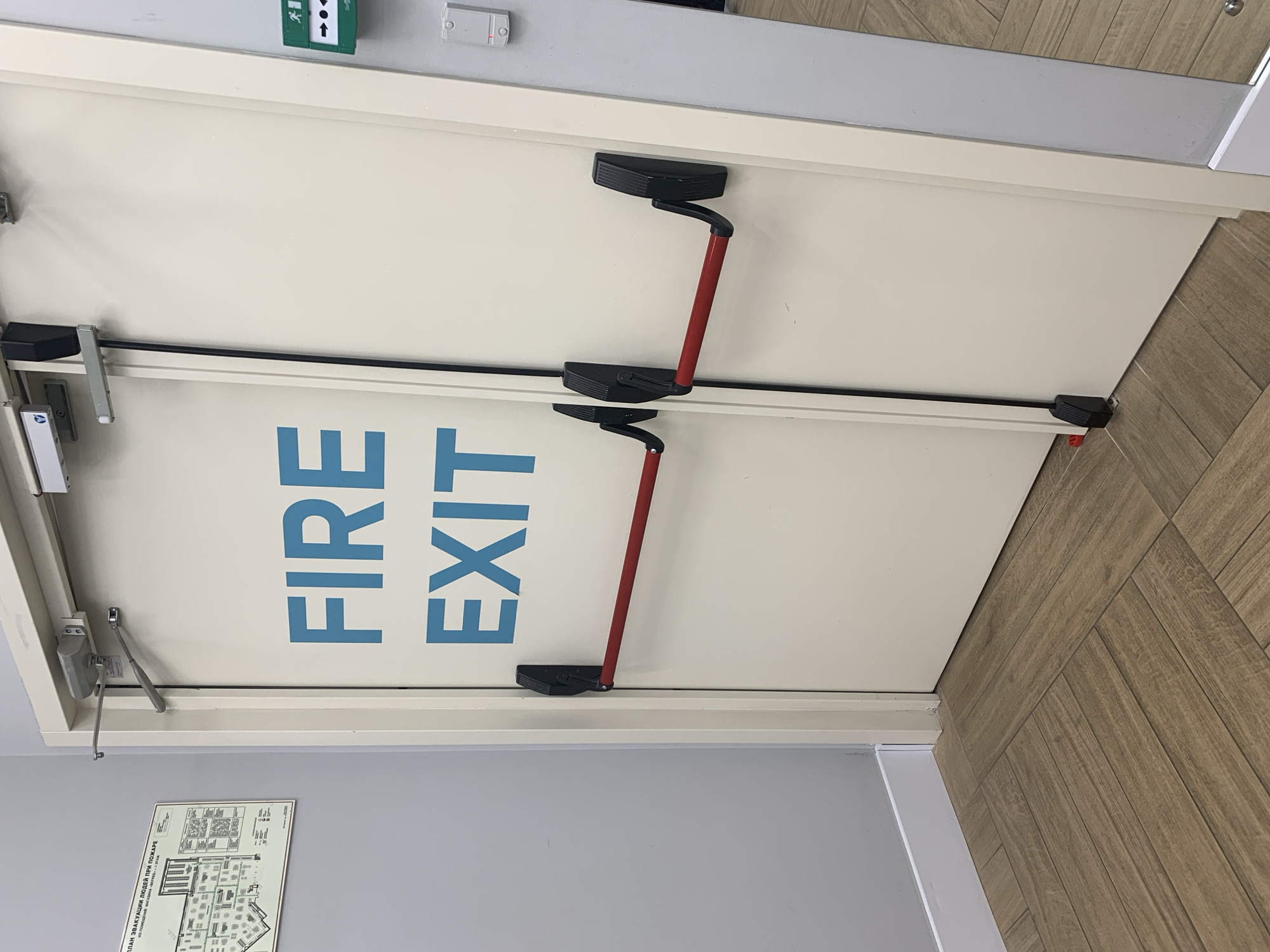The image shows a hallway with two double doors, likely an emergency exit. The doors are cream-colored and feature "FIRE EXIT" in large blue letters on the left door. Both doors have red push bars, which are commonly found on emergency exits for quick egress. To the left of the doors, a fire evacuation plan is posted on a light gray wall. The floor is covered with light brown wooden tiles laid in a herringbone pattern. The scene is well-lit, suggesting an indoor environment. No people are visible in the image, and no specific actions are taking place. The overall impression is one of a functional and well-maintained public space. Without further context, it is impossible to determine the specific location within Hrodna, Belarus.