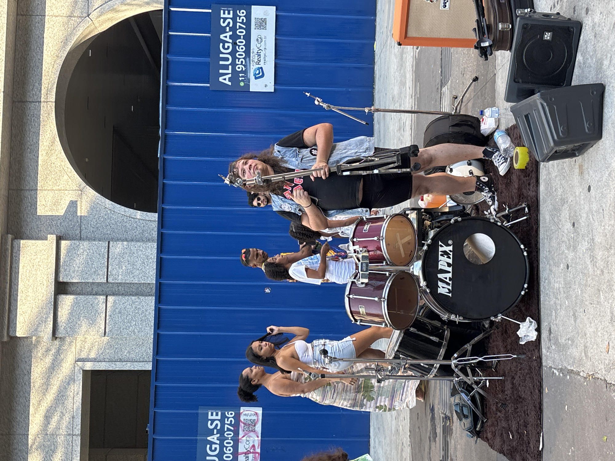 A street scene shows musicians performing on the sidewalk in São Paulo, Brazil. Three figures are visible in the performing group: two women, one in a floral dress and the other in white, both singing into microphone stands, and a male drummer behind them. Behind them is a second line of musicians: one male singing, the second one playing the trombone, and the third behind a drum set. They are performing in front of a blue, corrugated metal barrier. Advertisements are displayed on the barrier. A drum set, speakers, and other music equipment are set up on a brown mat on the sidewalk. A granite building with an arched entrance is to the left of the group.