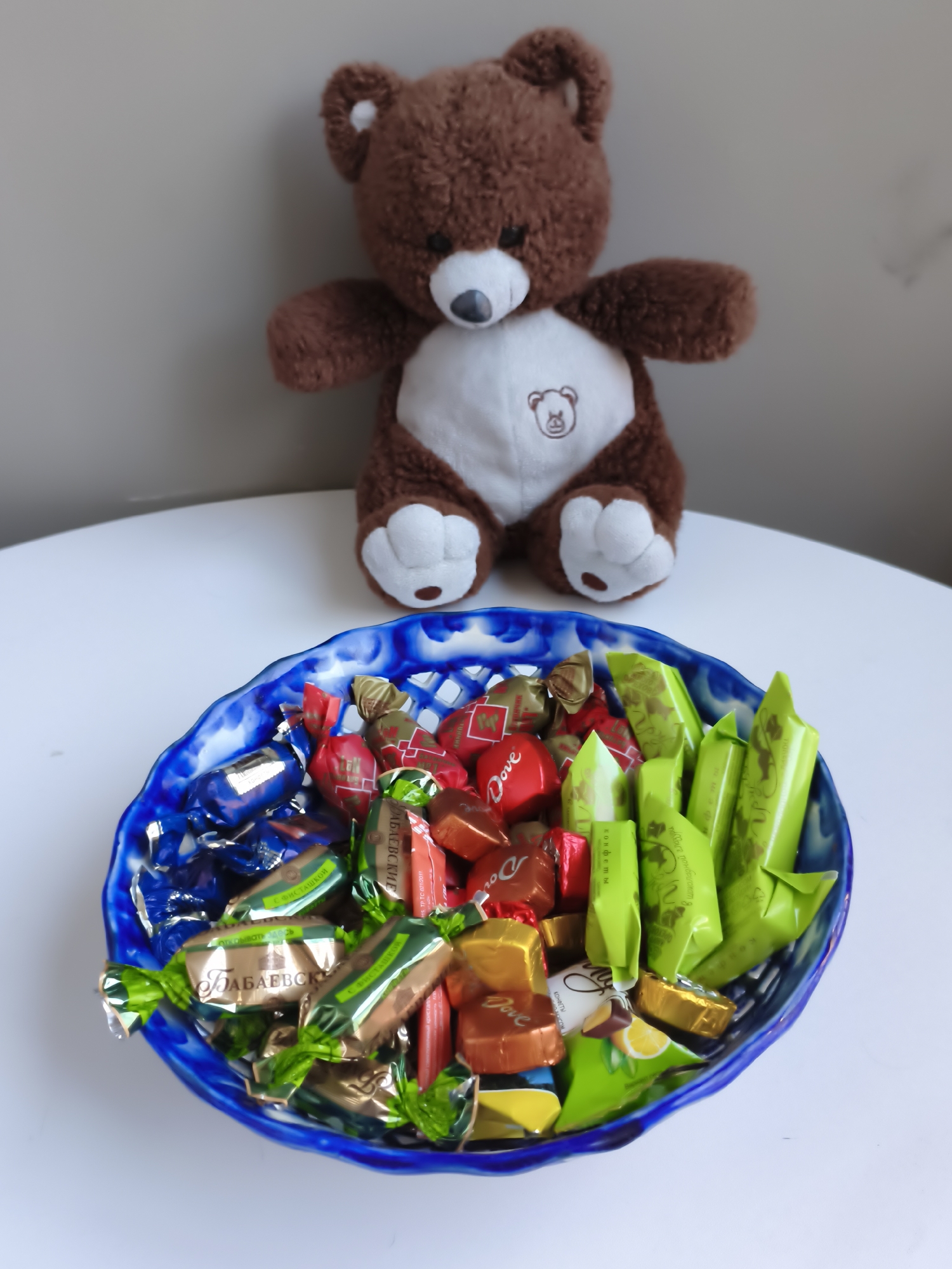 A brown teddy bear sits behind a blue and white patterned bowl overflowing with individually wrapped candies. The bowl is placed on a white circular surface, which is against a plain light gray wall. The teddy bear is plush, with black button eyes, a black nose, and a white belly with a small embroidered bear logo. The candies in the bowl are a variety of colors and wrappers, including red, blue, green, gold, and brown. Some of the wrappers have visible text and logos, such as "Dove." The candies are arranged in a haphazard pile within the bowl.