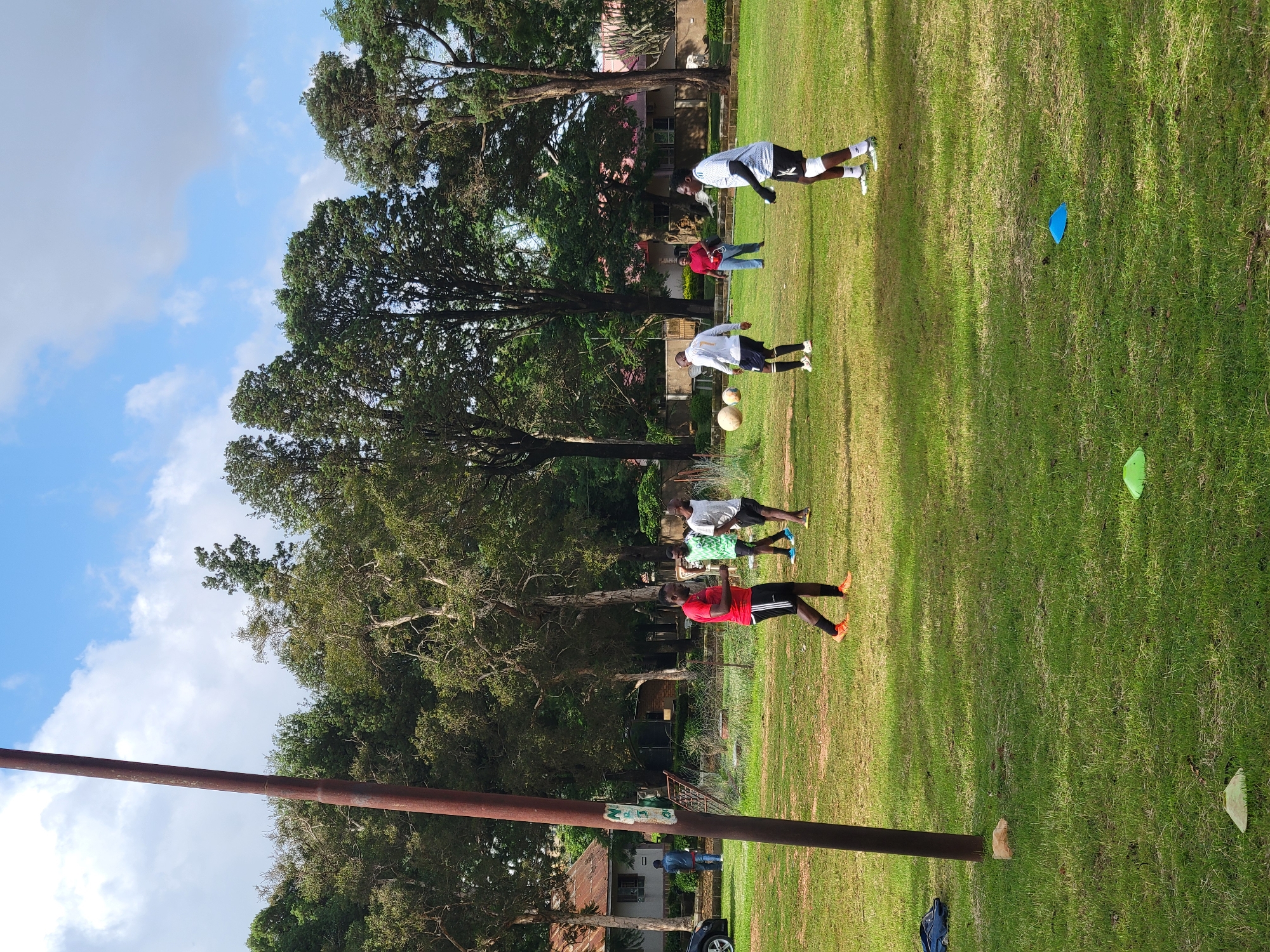 The image depicts a group of young men engaged in a football training session on a grassy field. The scene is outdoors, with lush green grass and trees bordering the field. The sky is partly cloudy, suggesting a pleasant day. The time appears to be daytime, given the clear shadows cast on the grass.
The subjects are primarily the young men, dressed in athletic wear. Several are actively participating in drills, including one in a red jersey and black shorts, another in a white t-shirt and black shorts, and others in similar sportswear. Two footballs are visible on the field, indicating the nature of the activity. There are also training cones scattered on the grass, one blue and one green, further suggesting a structured training exercise. In the background, buildings and houses are visible, peeking through the trees, hinting at an urban or semi-urban environment. A tall, rusty pole, possibly part of a goalpost or other sports equipment, runs vertically along the left side of the image. A dark blue sports bag is also visible on the ground near the pole.
The overall impression is one of focused athletic activity in a communal setting. The participants appear determined and engaged in their training. The setting is typical of an outdoor sports field, likely in Jos, Nigeria, given the location context.