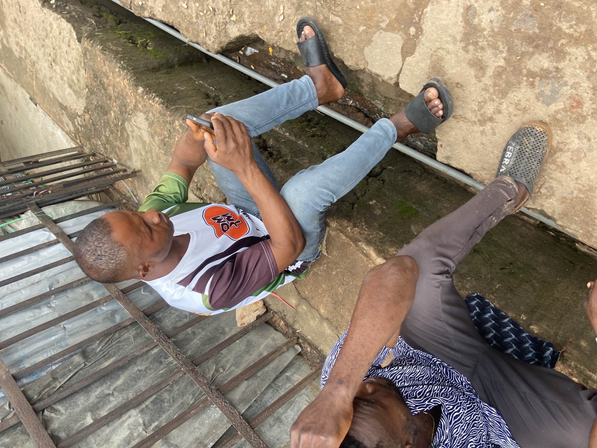 Two men are seated on a concrete ledge in Ogidi, Nigeria. The man on the right, wearing a white shirt with colorful sleeves and blue jeans, is holding a smartphone and looking down at it. He has short hair and is wearing black slides. The man on the left, wearing a patterned shirt and grey pants, has his head in his hands and appears to be distressed. He is wearing perforated grey shoes. The environment includes a metal fence or barrier behind the men, and a narrow, moss-lined trench to their right. The ground is a rough concrete surface.