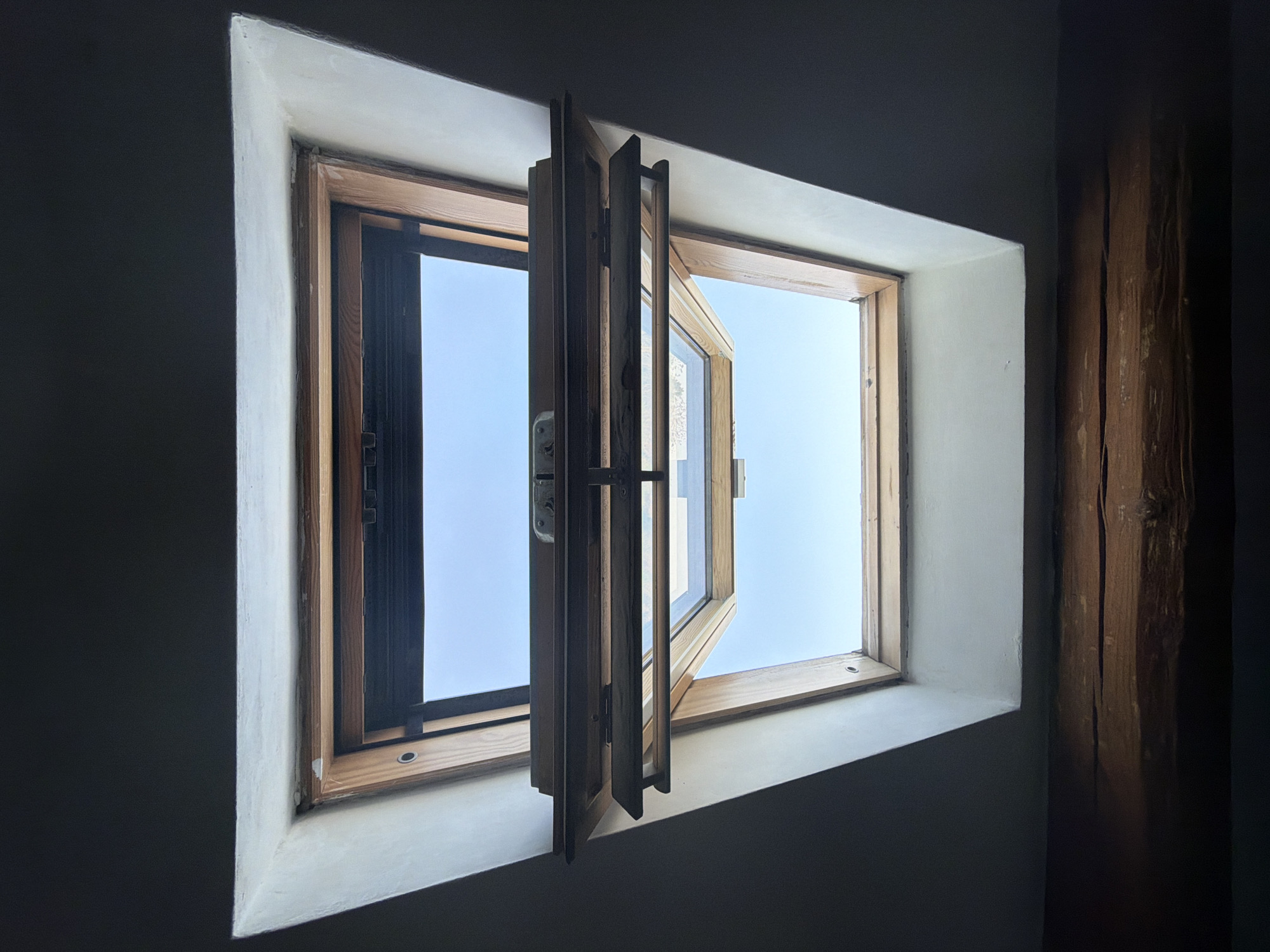 An eye-level, interior shot depicts a partially-open, wooden window frame set within a pale, plastered wall. The window reveals a clear, blue sky. The left pane of the window is partially closed, while the right pane is open to a greater degree. The photo is shot in low light, with the surrounding walls and ceiling predominantly dark. A portion of a distressed wooden beam is visible on the right-hand side of the frame. The image provides no information to indicate it was taken in Milan, Italy. No people or interactions are discernible.