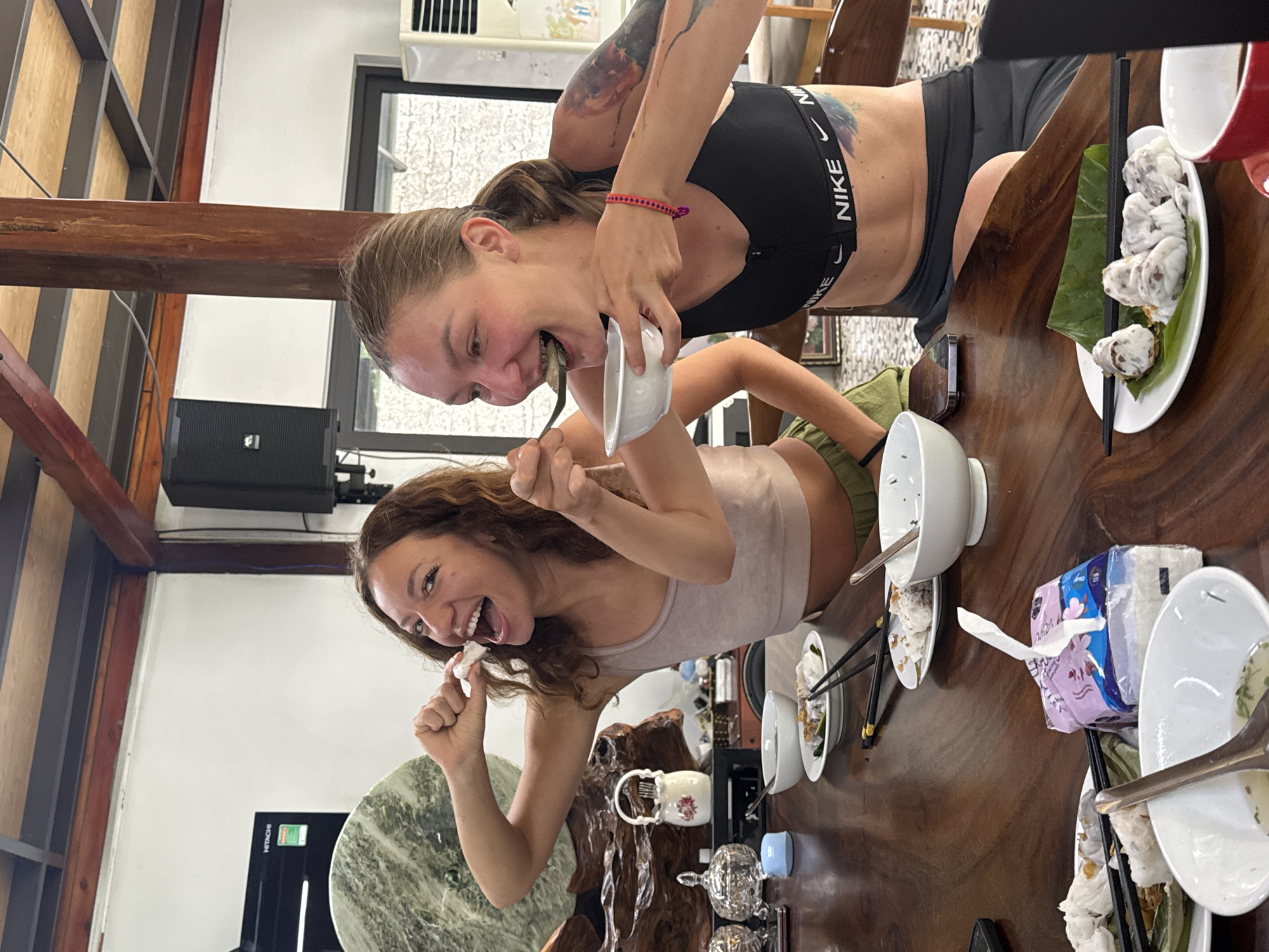The scene depicts two women interacting around a large wooden table covered with plates, bowls, and utensils, suggesting a meal or dining experience. The woman closer to the camera is holding food in her hand and is smiling broadly, while the other woman is eating directly from a bowl, also with a joyful expression.
The table setting indicates a potential Asian dining experience, with chopsticks visible among the utensils. There are dishes containing what appear to be dumplings, rice, and other food items.
The environment is indoors, with wooden beams and a stone table visible in the background. A black speaker is mounted on the wall and there is an air conditioning unit visible in the background.
Based on the information provided, the location is likely in Hà Giang 1 Ward, Vietnam.