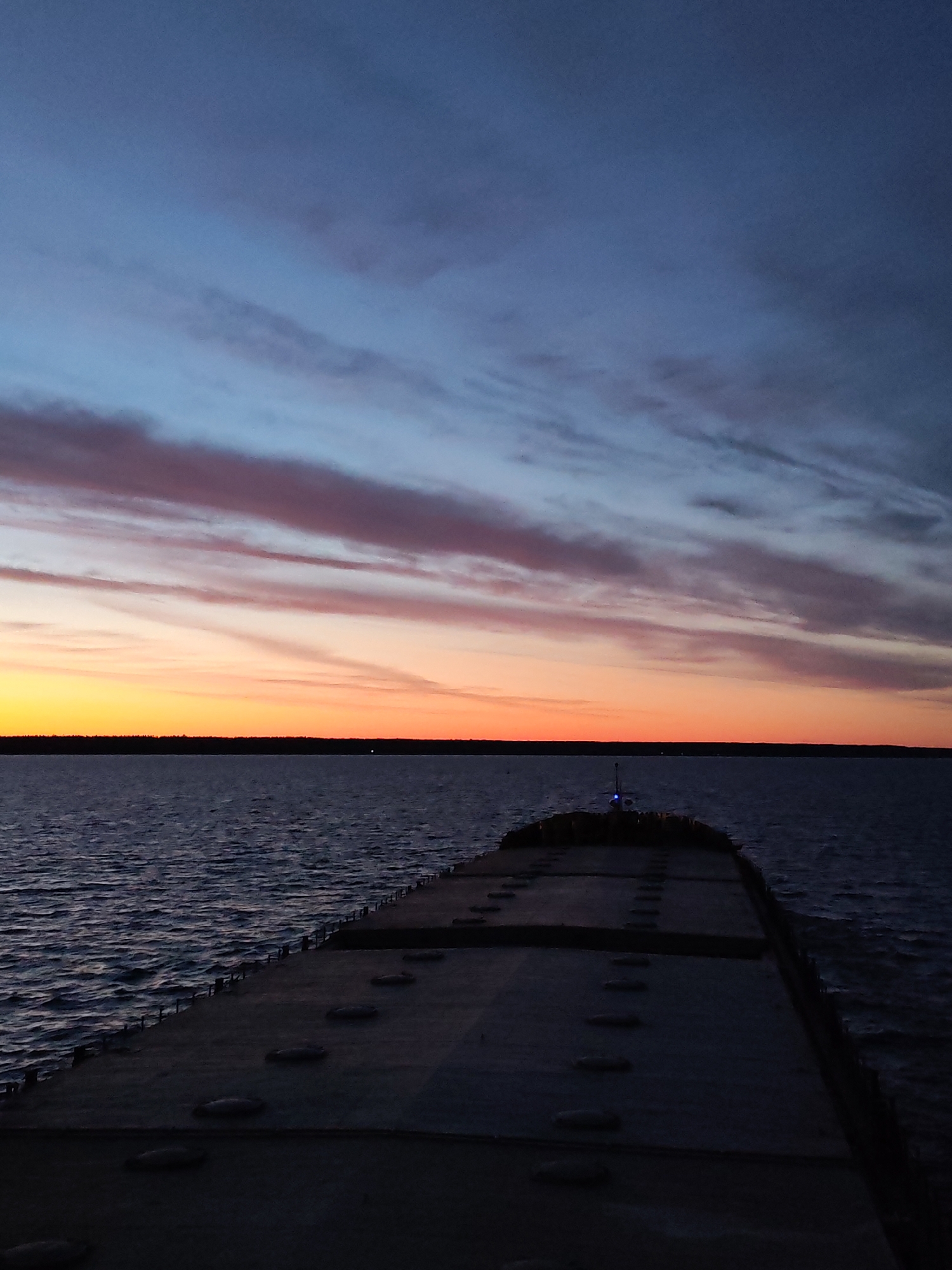 The image shows a view from the deck of a vessel at dusk. The deck is gray and features numerous evenly spaced, round hatches. The dark water stretches out to meet the horizon, where a dark treeline is visible. Above the horizon, the sky displays a gradient of colors from orange to pink to deep blue. Clouds are present in horizontal bands. A small light is visible at the end of the deck.