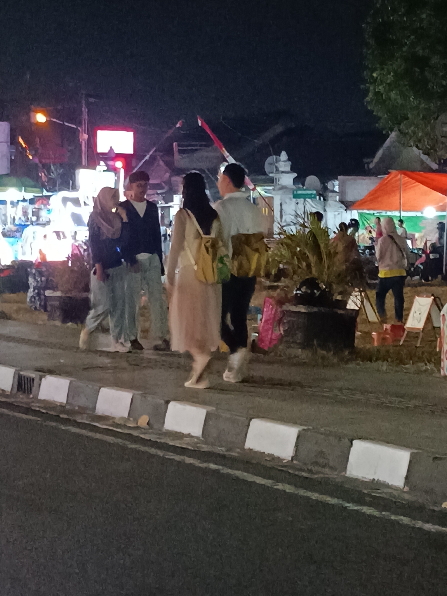 At night, several people are walking along a street in Yogyakarta, Indonesia, with various market stalls and tents set up on the side of the road. A group of three individuals is in the foreground: a woman in a headscarf and a man in a dark jacket and light-colored pants are walking arm-in-arm, while another woman in a light-colored dress with a backpack walks ahead of them. Further back, other individuals are visible browsing the stalls. The scene is illuminated by artificial lights from the market and streetlights. The road is paved, with a concrete curb and white markings. Trees and some buildings are visible in the background.