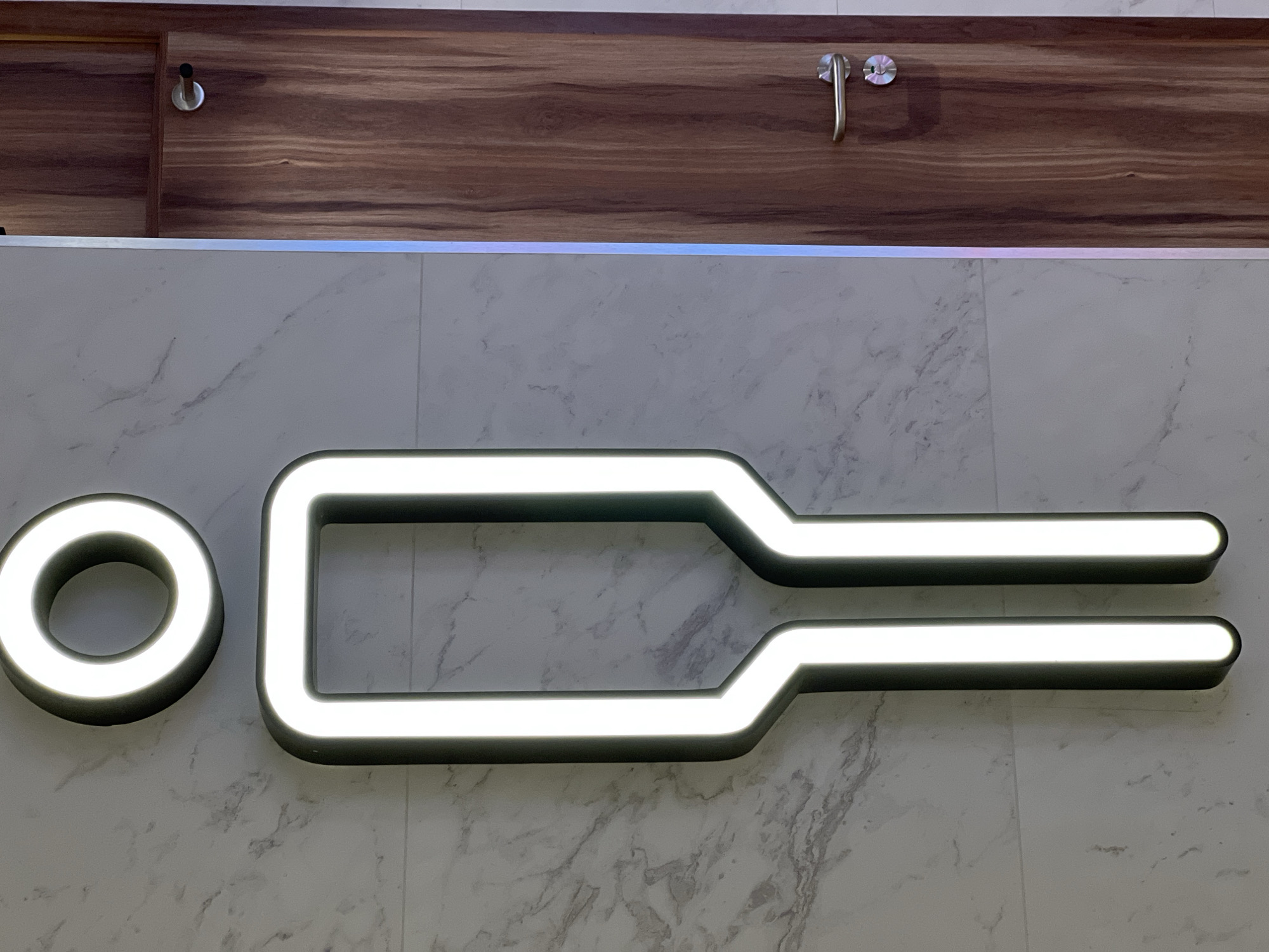 A modern, illuminated restroom sign for men is mounted on a marble-tiled wall next to a wooden door with a silver handle. The sign is a stylized, outlined figure of a person with a circular head and a rectangular body that tapers to two legs. The outline is illuminated with bright white light. The background wall is made of large marble tiles with a grey veined pattern. To the right of the sign, a wooden door is visible, featuring a natural wood grain texture and a silver door handle and lock. A thin, vertical strip of iridescent light runs between the wall and the door. The image is taken from a slightly low angle, looking up at the sign and door. The overall setting suggests a clean and contemporary public restroom. The image does not provide specific information about the city of Derry/Londonderry, United Kingdom.