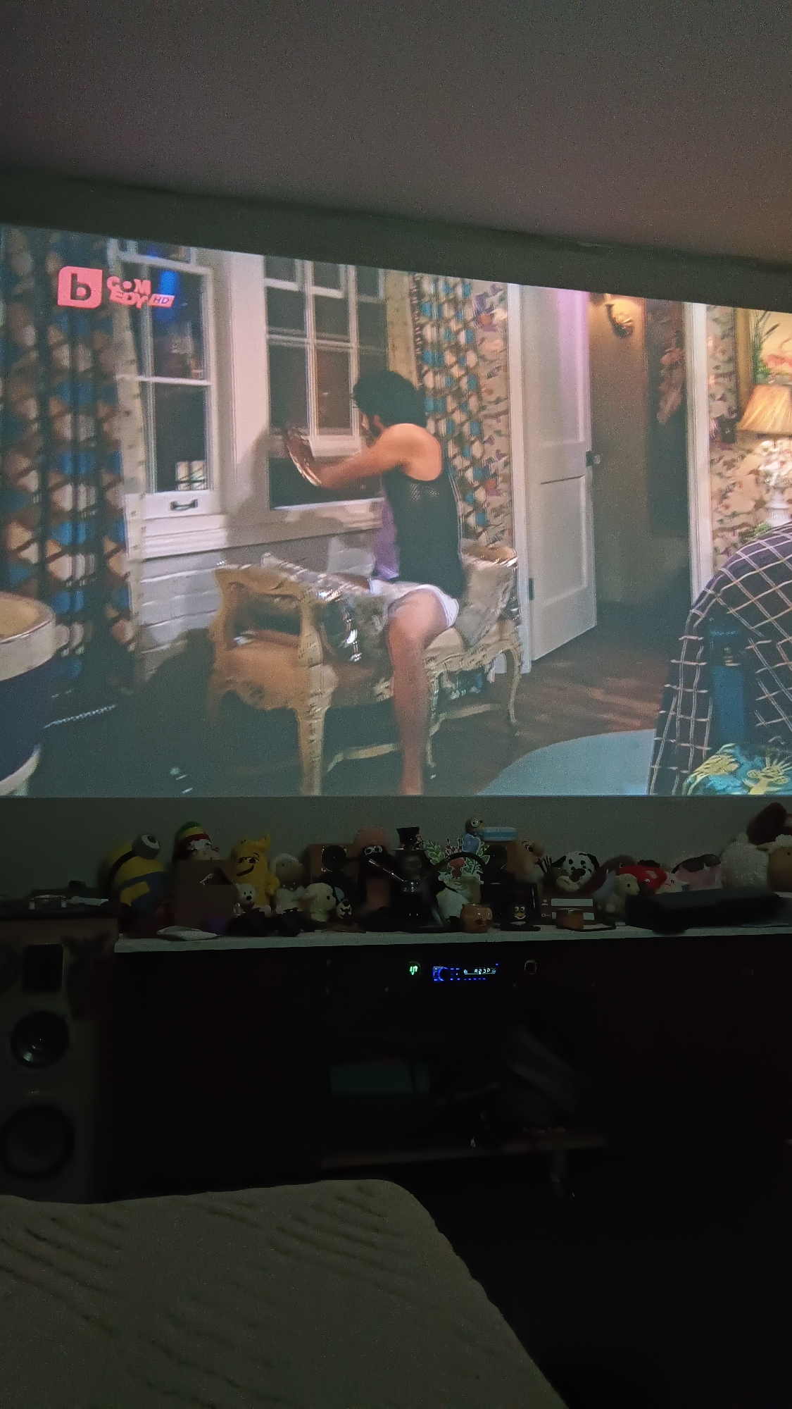 The image displays a projected video on a large screen in a dimly lit room, with a shelf of objects and an electronic device beneath it.
The projected video features an indoor scene with a person, viewed from the back and side, leaning or sitting on an ornate, gold-colored chair. The person wears a black tank top and white underwear or shorts, and is seen reaching towards a white-paned window, holding a circular, reflective object. The room in the video is decorated with patterned curtains, a white door, a wall sconce, and a table lamp. The channel logo "b COMEDY HD" is visible in the top left corner of the projection.
Below the screen, a shelf holds a diverse collection of stuffed animals and figurines. Identifiable items include a yellow Minion, a red-capped character, a yellow plush toy, several small animal plushies (some white, some brown), a hookah pipe, a figure with a large red headdress, and a small orange pumpkin decoration.
In the lower part of the image, an electronic receiver with a blue digital display (showing "EC" and other unreadable characters) is visible. A speaker is partially seen to the left. The immediate foreground is a textured, light-colored surface, possibly a blanket or mattress, suggesting the photo was taken from a comfortable position in the dark room, likely at night.