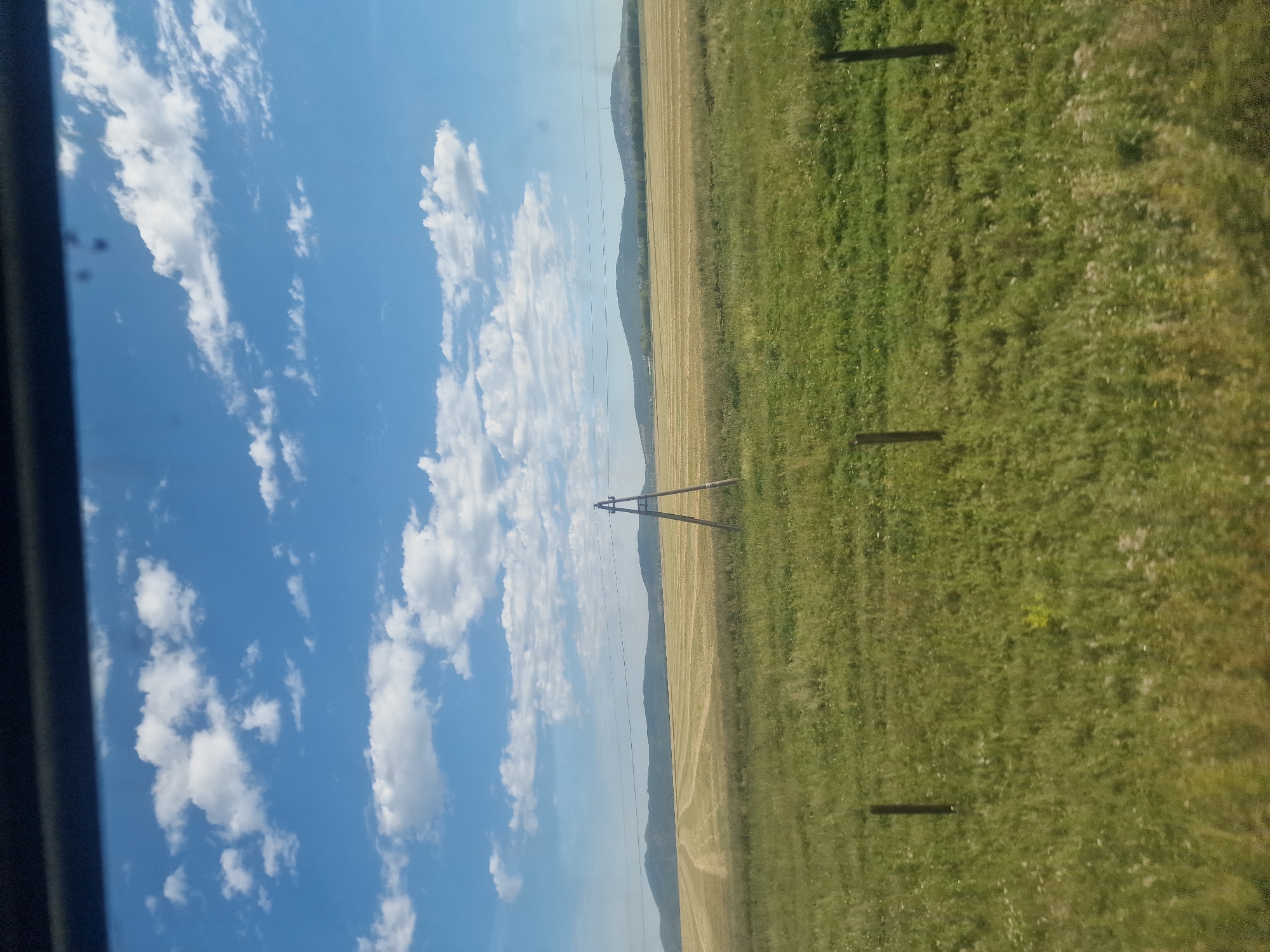 A wide-angle outdoor shot depicts a rural landscape under a partly cloudy blue sky. In the foreground, a grassy field with scattered small white flowers and a few fence posts is visible. A stark wooden A-frame structure stands in the middle ground, likely a utility pole or support for wires, with power lines extending horizontally across the scene. Beyond this, a vast, harvested field of light brown stubble stretches towards rolling hills in the background. The sky is a vibrant blue with scattered, fluffy white clouds. The scene suggests a rural setting within the Абылайхан ауылдық округі, Kazakhstan, characterized by agriculture and open spaces.