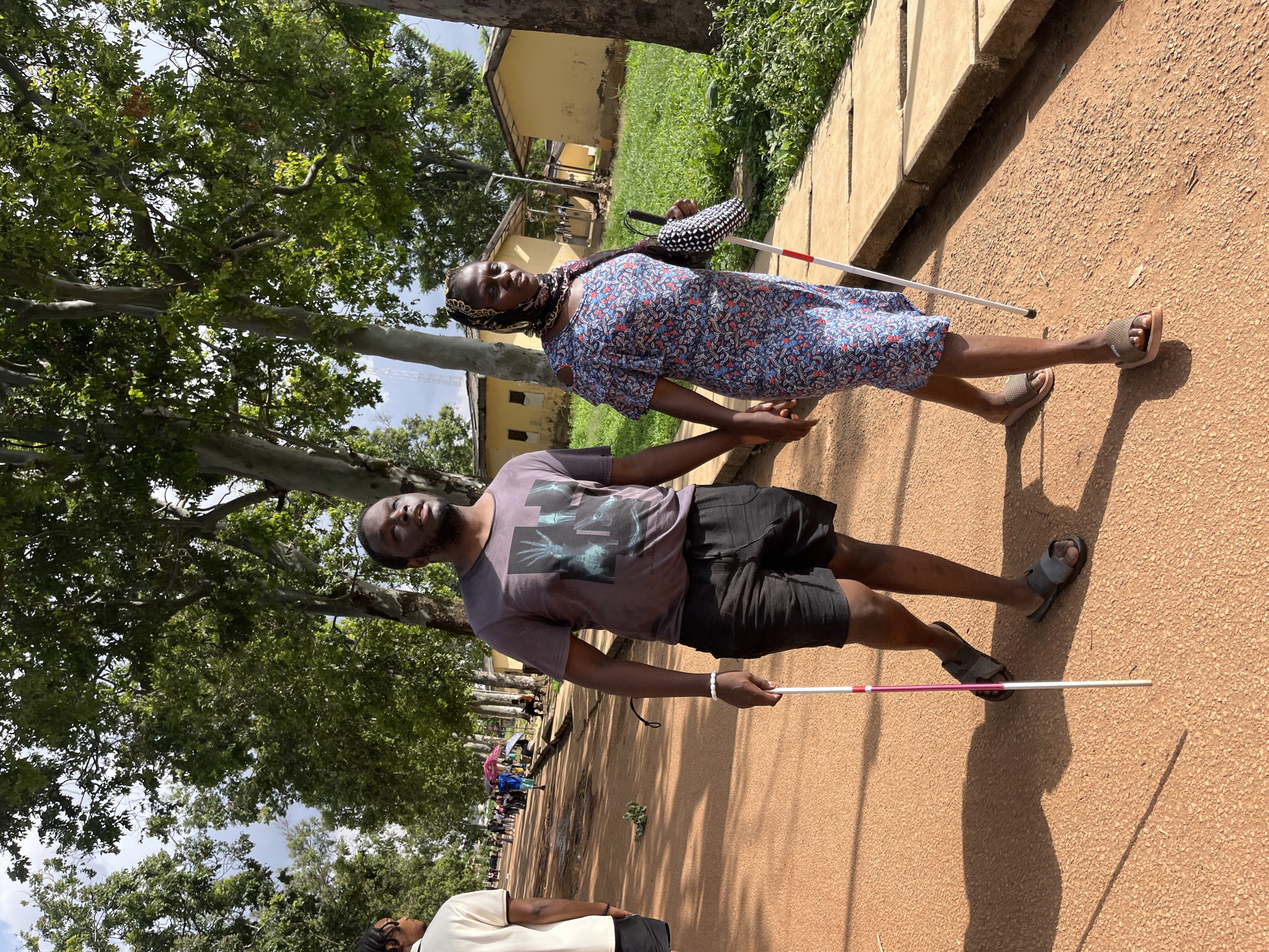 A man and a woman are walking outdoors, holding hands. The woman is on the left, wearing a floral-print dress and a headscarf. She is holding a white cane with a red tip in her right hand and a patterned bag in her left. The man is on the right, wearing a grey t-shirt with a graphic print and black shorts. He is holding a white cane with a red tip in his right hand. They are walking on a reddish-brown path, with trees and buildings in the background. The scene appears to be set in Jos, Nigeria. The sun is shining, casting shadows, indicating it is daytime. The overall mood seems casual and possibly educational or supportive, given the canes and the way they are walking together. There is no visible text in the image.