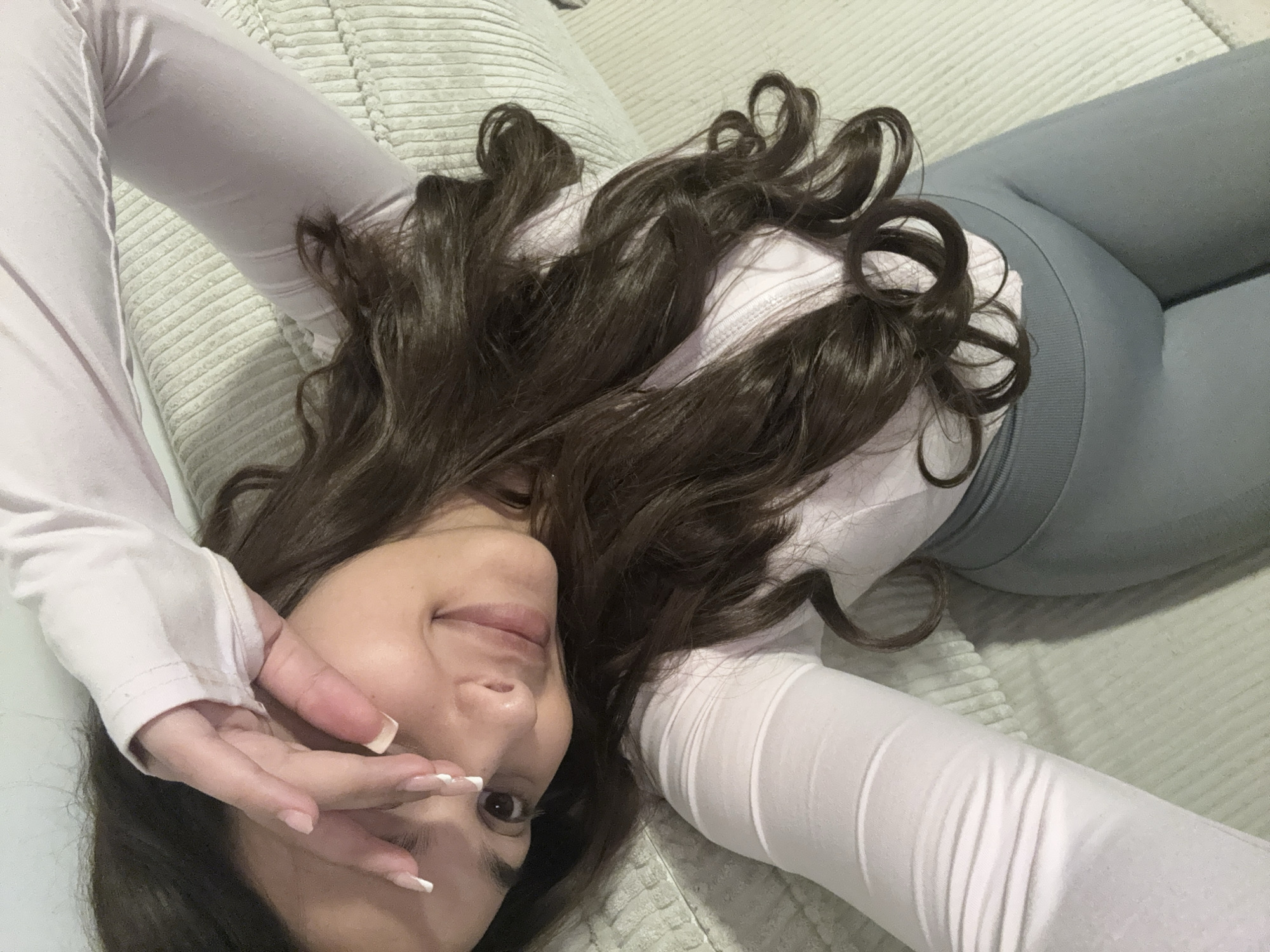 A woman with long brown curly hair is lying on her back on a light-colored, textured surface, possibly a couch or bed. She wears a light-colored, long-sleeved top and grey leggings. Her arms are outstretched, and one hand is partially covering her face. She is smiling. The location is unknown and there is no indication of the city of Mönchengladbach, Germany.