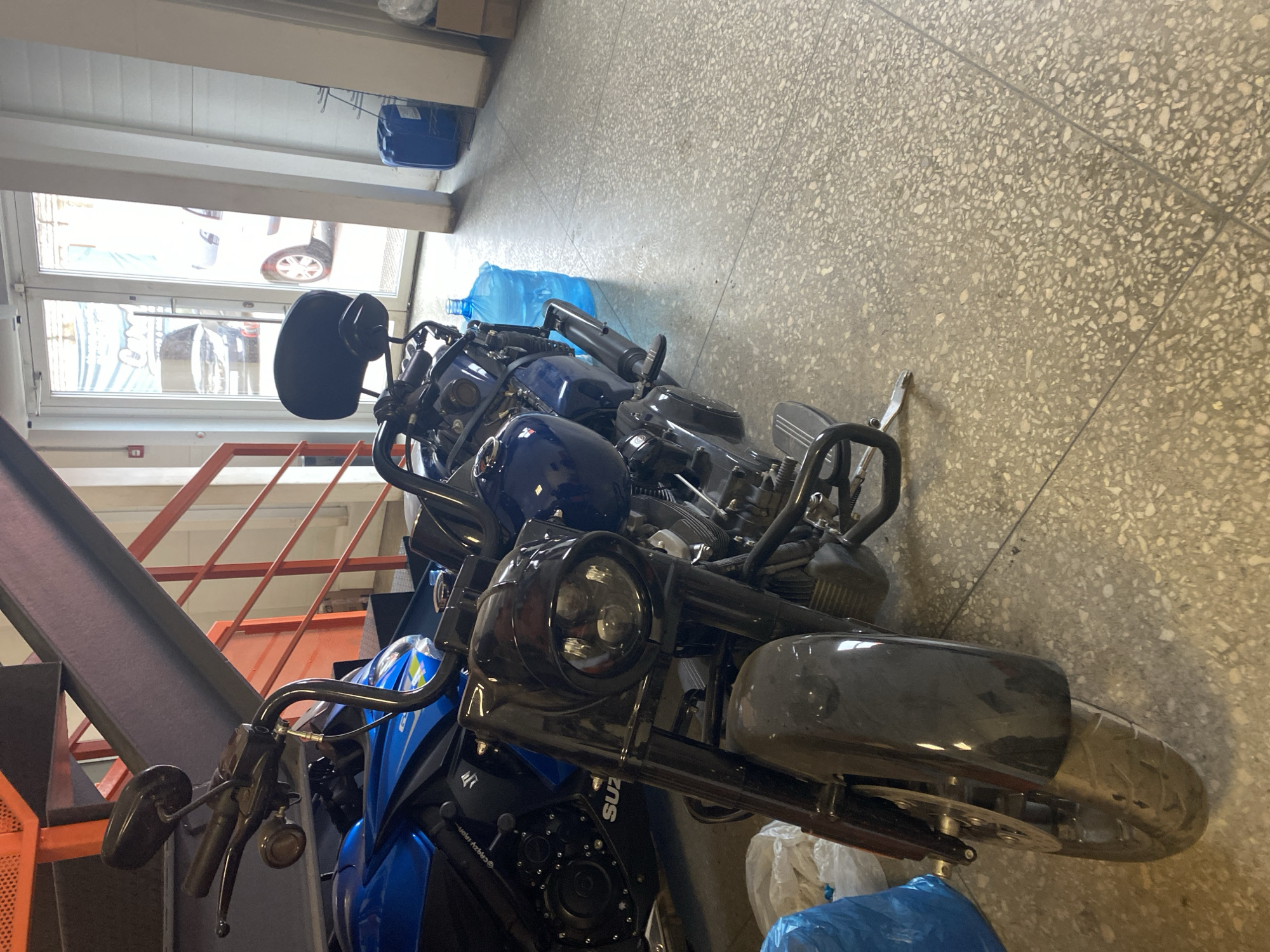 The image depicts a close-up view of several motorcycles parked indoors, potentially a storage area or garage, in the city of Svkh Voronezhskiy, Russia. The motorcycles are predominantly blue and black. Other details:
* The setting appears to be a commercial or industrial building with a tiled floor.
* There is a metal staircase with orange railings visible on the left side of the frame.
* A window is visible in the background, revealing a view of a parked car.
* Several blue plastic bags are scattered around the motorcycles.