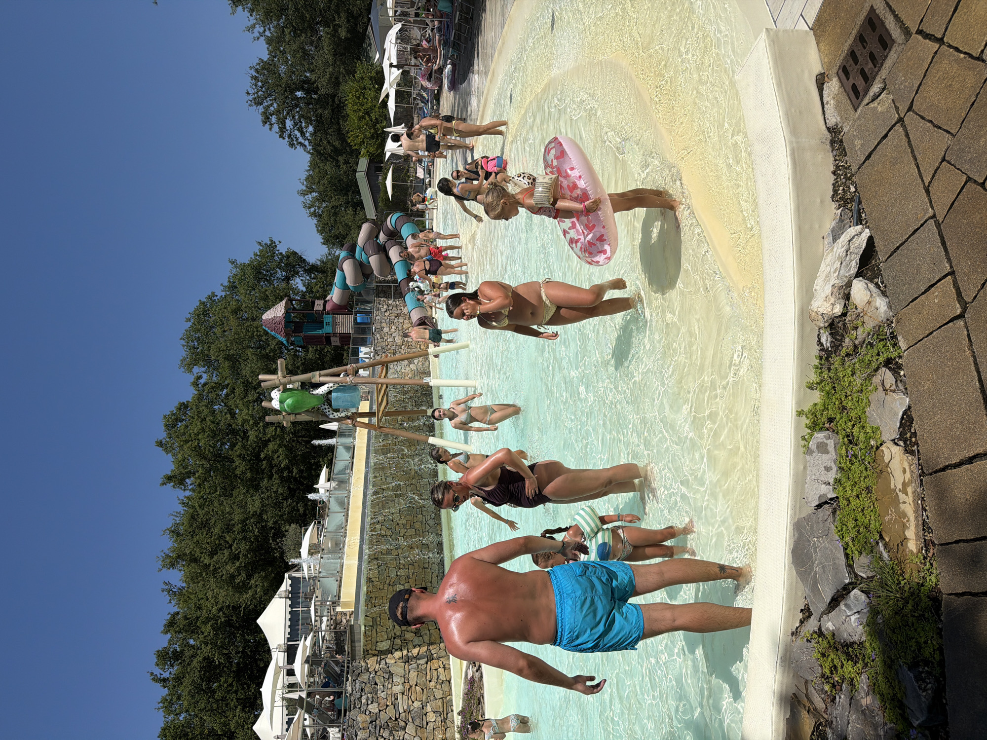 A group of people are enjoying a sunny day at a water park. The scene is a shallow pool with clear, bright water, with a sandy or light-colored bottom. Several individuals are standing or wading in the water. In the foreground, a man wearing blue swim trunks and a black baseball cap is facing away from the camera, his back to the viewer. To his left, a young girl in a swimsuit and a striped shirt stands with a pink inflatable ring patterned with red hearts. Further into the pool, other people are visible, some in swimsuits, appearing relaxed and enjoying the water.
Behind the pool, a large water slide structure with colorful segmented tubes is visible. Beyond that, lush green trees form a backdrop under a clear blue sky. There are also white umbrellas and lounge chairs set up, suggesting a recreational area. The image is captured in daylight, likely midday, given the brightness and the shadows cast. The overall atmosphere appears to be one of leisure and fun, characteristic of a summer day at a water park. There is no visible text in the image. The location context provided is Cavriglia, Italy, which aligns with the warm, sunny weather and outdoor recreational setting.