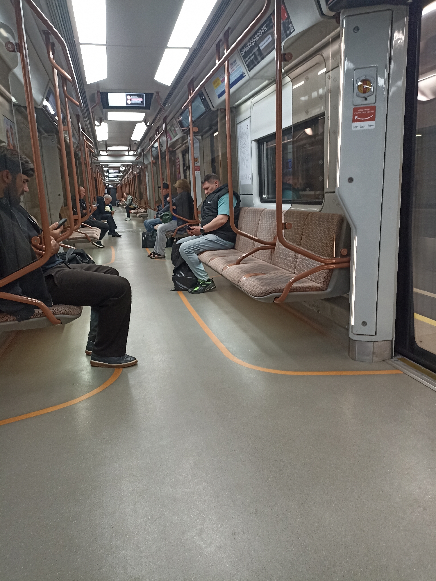 The image shows the interior of a Moscow Metro carriage. Several passengers are seated on brown and beige upholstered benches. The floor is a neutral gray with an orange line marking a safety zone. The walls are a light gray, and the ceiling has bright white fluorescent lighting panels. On the right side of the image, a grey vertical column with a red and yellow emergency button and a sign with Russian text is visible. Passengers are dressed in casual clothing, with some looking at their phones. The overall scene depicts a typical urban public transportation environment.
