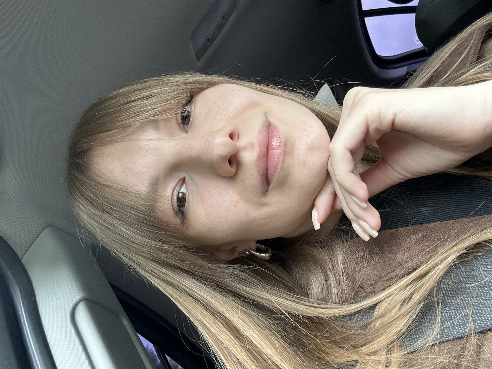 A young woman with long blonde hair and light skin is taking a selfie in what appears to be a car. She is holding her hand to her chin. She is wearing silver hoop earrings. The location of the photograph is not indicated within the image, so its being taken in Lukintsy, Russia cannot be verified from the visual data.