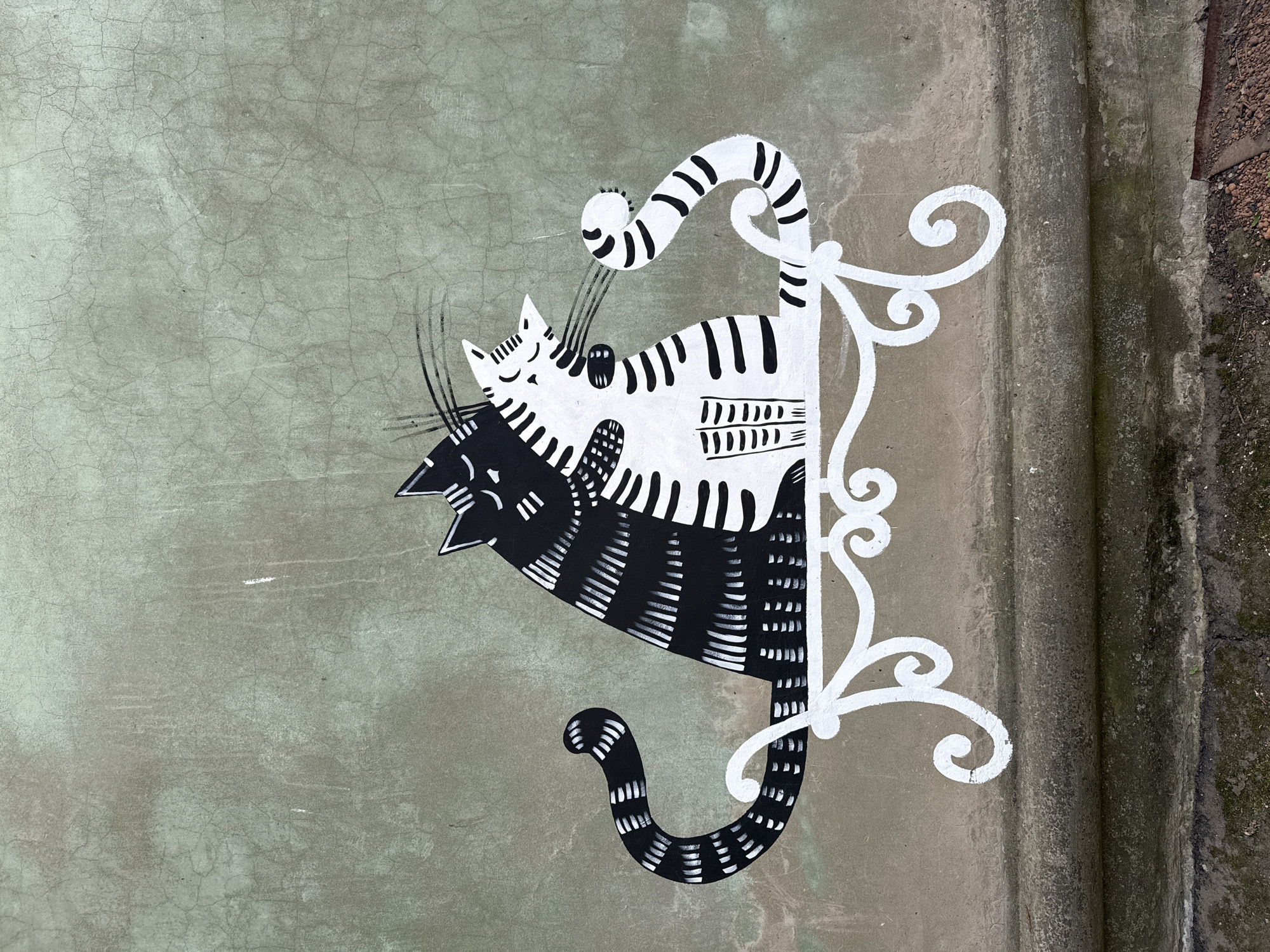 The image shows a black and white mural of two cats, one on top of the other, on a weathered concrete wall. The upper cat is mostly white with black stripes, while the lower cat is mostly black with white stripes. A decorative white element, resembling a stylized support bracket or frame, is painted adjacent to the cats. The wall is cracked and textured with a light gray-green hue. The edge of the wall is visible on the right, showing darker, possibly mossy, areas and hints of brown pebbles or dirt. The photo was taken outdoors in Votkinsk, Russia.