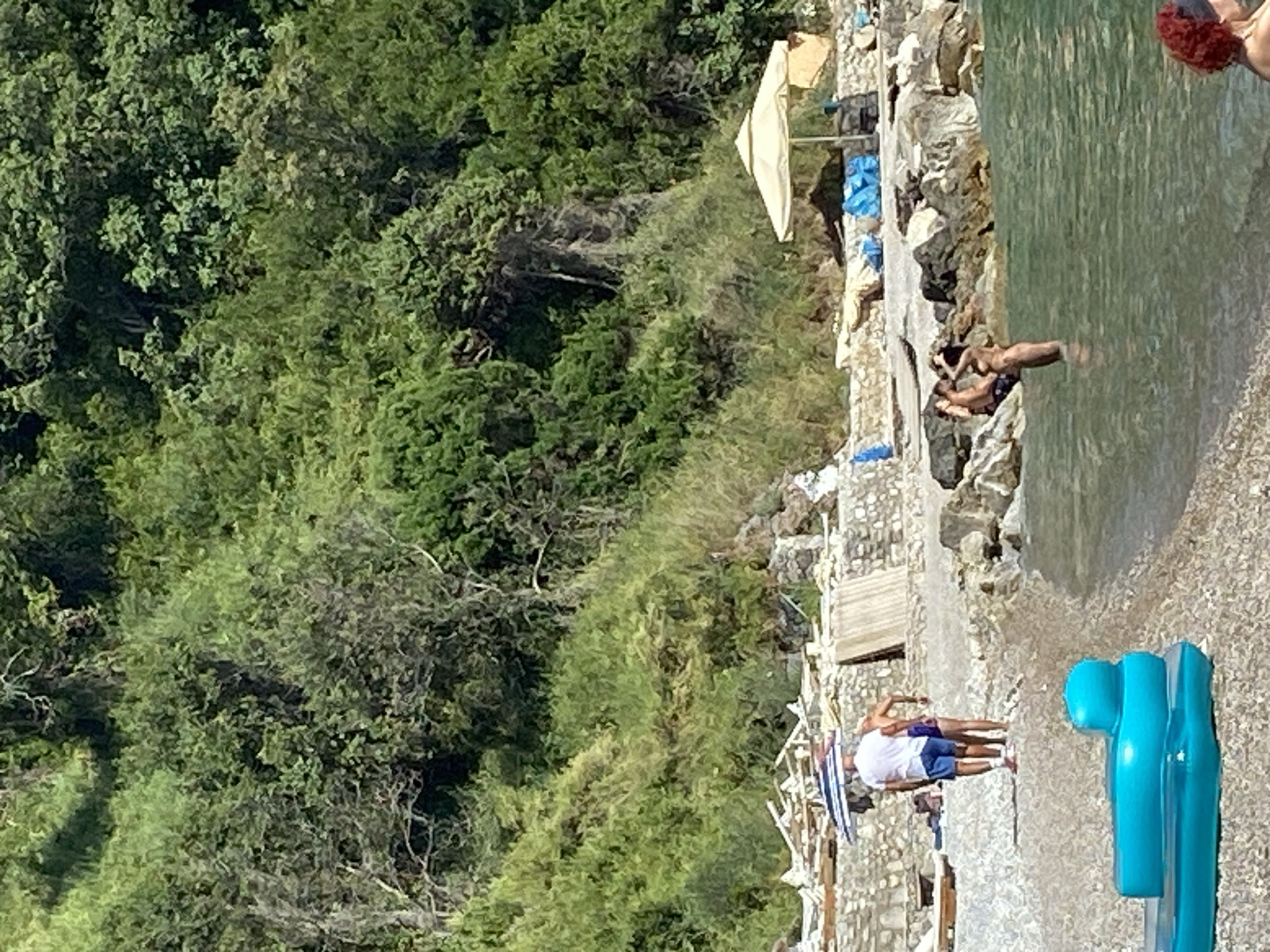 The image depicts a sunny day at a rocky beach in Prijevor, Montenegro. The background is dominated by a lush green hillside covered in dense vegetation and trees. In the middle ground, a stone wall separates the beach from the hillside, with some wooden structures and lounge chairs visible. A large, light-colored umbrella provides shade.
On the beach, a couple walks together under a striped umbrella, with the man wearing a white t-shirt and blue shorts, and the woman in a purple swimsuit. A blue inflatable float is positioned on the pebbled shore in the foreground.
In the water, a couple is interacting, with one person partially submerged. The water is clear and appears shallow near the shore. The overall scene conveys a relaxed atmosphere of a summer day at the beach.