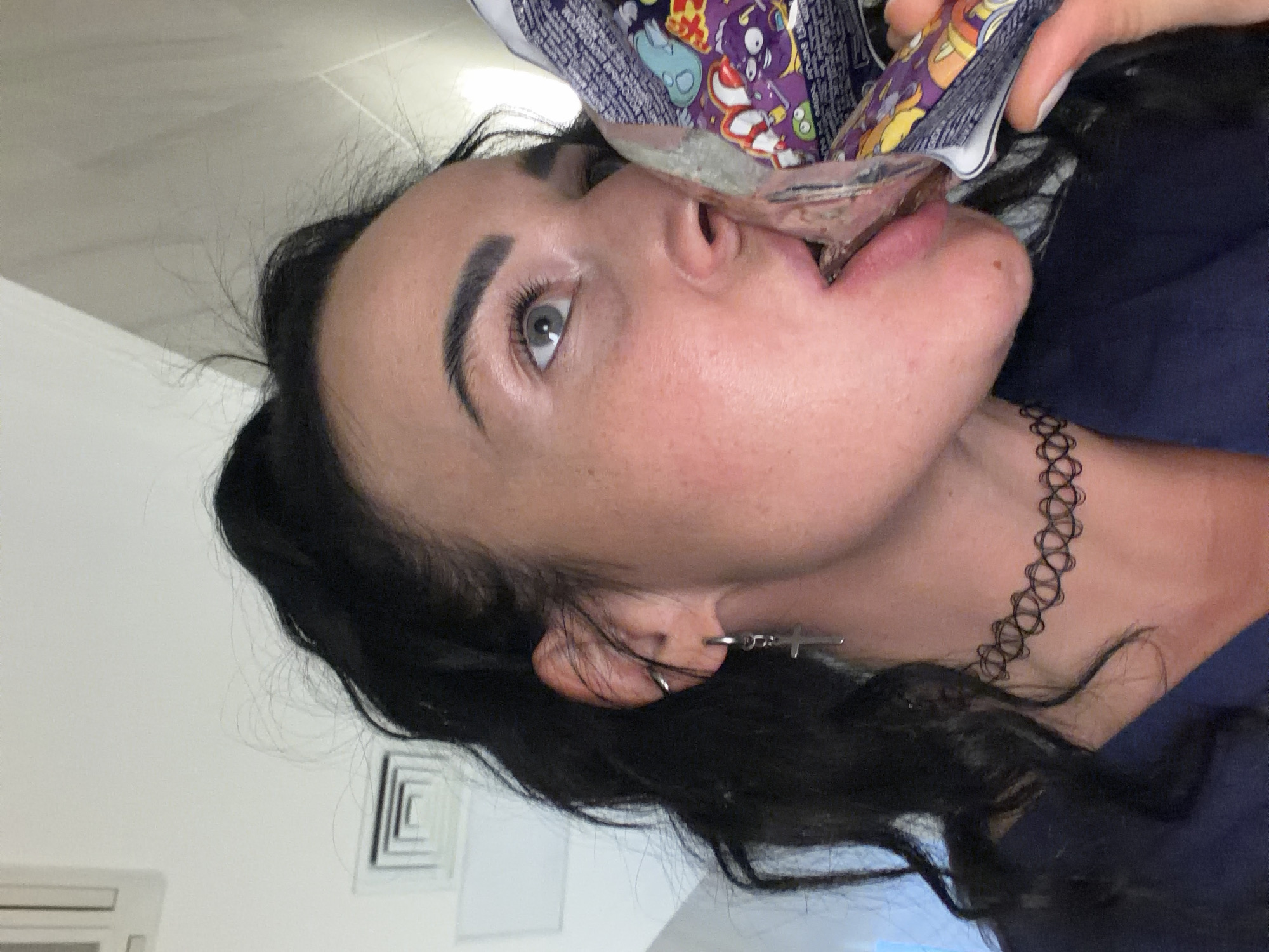 A woman with dark hair pulled back in a ponytail is eating from a foil bag of some kind of snack. She has a black choker necklace and a silver cross earring in her left ear. The background shows a white wall and a vent. The location is not discernable from the image.