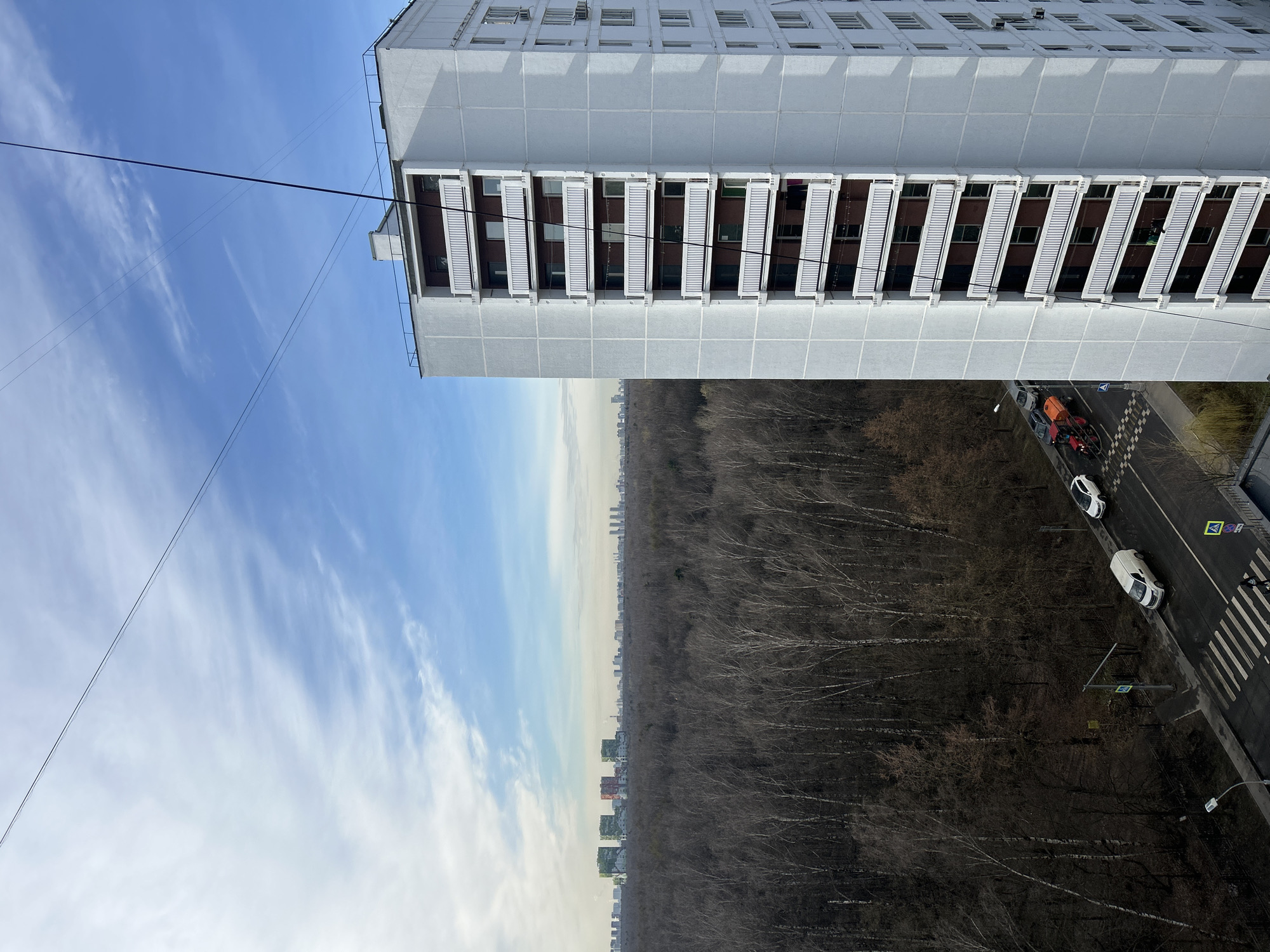 The image shows an elevated view, possibly from a high-rise building in Moscow, Russia. A tall, rectangular apartment building dominates the upper portion of the frame. It is gray and white with multiple rows of windows and what appear to be external sun shades. A power line is visible extending into the sky.
Below the building is a dense forest of bare trees, indicating a cold season, likely late fall or winter. A road runs parallel to the forest edge, with a crosswalk. A few cars are parked or driving on the road. A city skyline is faintly visible on the horizon beyond the forest.