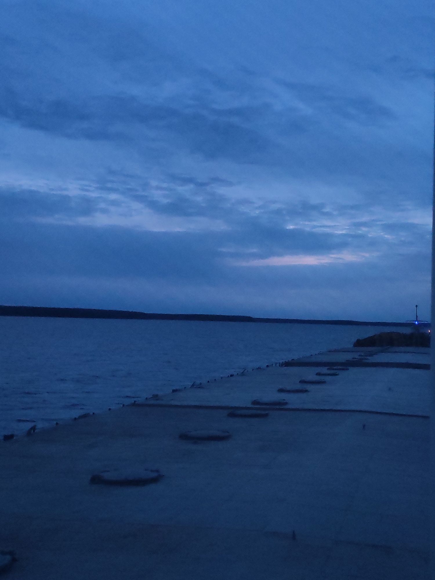 The image depicts a waterfront view at dusk or dawn. The sky is predominantly blue with cloud cover. Below the sky, a dark treeline or distant landmass stretches across the horizon. A body of water fills the middle ground, reflecting the dim light from the sky. In the foreground, a concrete pier or walkway extends into the water. There are several recessed circular shapes evenly spaced on the surface of the pier. A structure with a blue light is visible on the pier in the distance. There are no discernible people or interactions captured in the scene. Based on location information this is likely the Volga River waterfront area of Topornya, Russia.