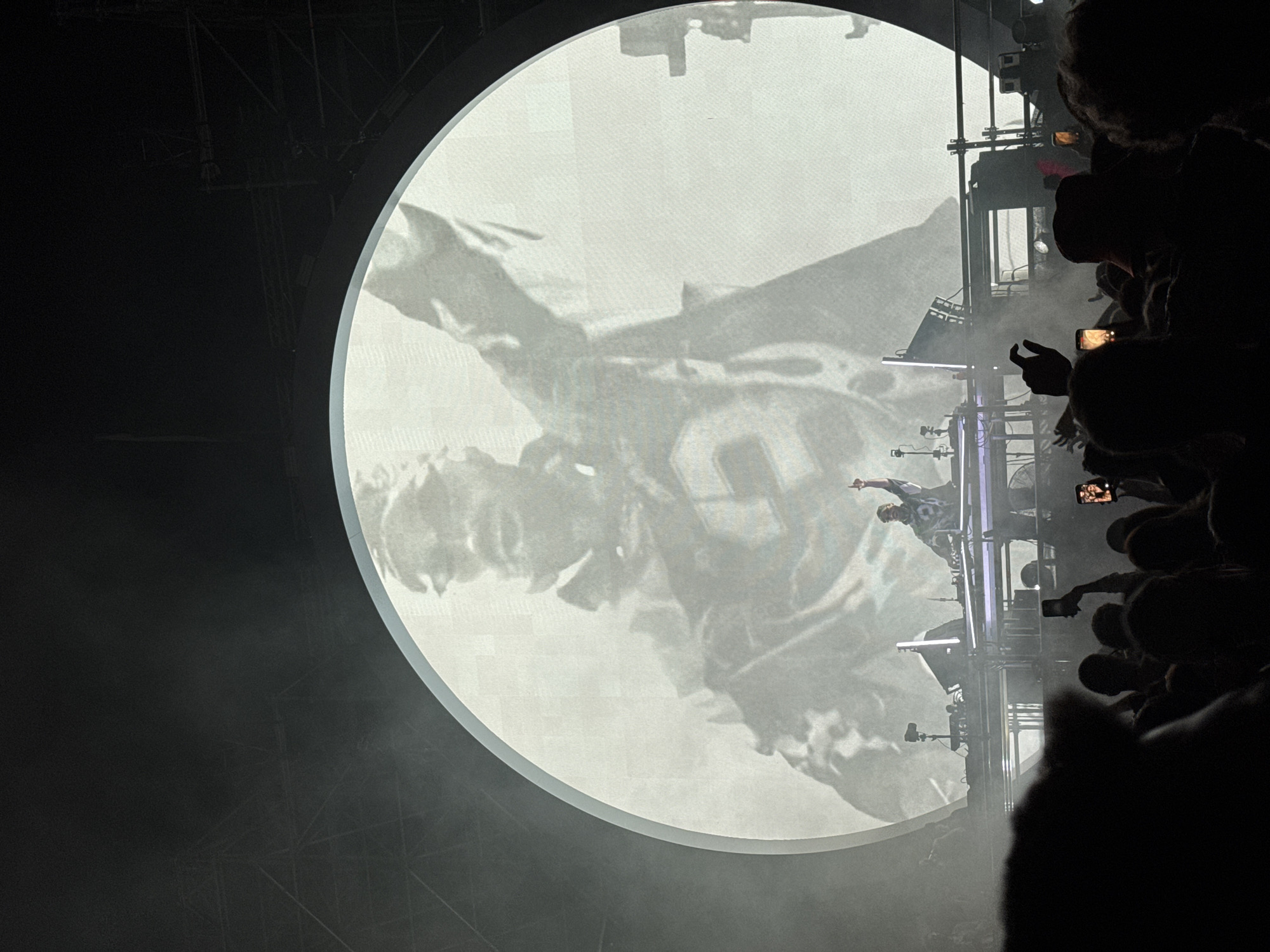 The image captures a live music performance on a stage with a large circular screen in the background displaying a black and white graphic of a person wearing a baseball cap and a jacket. A DJ is standing behind a console on the stage, illuminated by stage lights and a projection of the graphic. The DJ is seen with one hand raised. The stage is equipped with speakers and other audio equipment. In the foreground, a crowd of concert-goers is visible, many holding up their mobile phones, presumably to record or photograph the event. The atmosphere appears to be energetic, with smoke or fog effects present on the stage. The scene suggests a large-scale music event, possibly a concert or festival. No specific location in Sofia, Bulgaria is identifiable from the visual information provided.