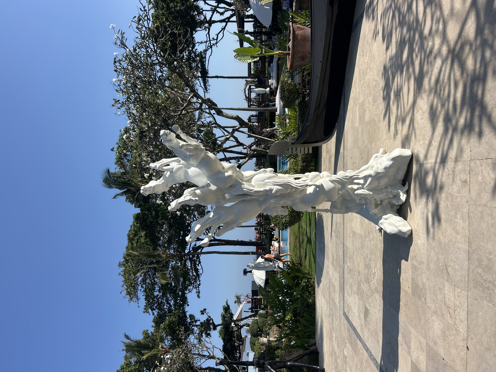 In North Kuta, Indonesia, a white abstract sculpture of horses in motion stands prominently on a paved area. To the right of the sculpture, a black gondola boat is positioned, partially obscured by the sculpture and a large potted plant. Beyond the paved area, a swimming pool is visible, with lounge chairs and umbrellas scattered around it. Lush green trees and foliage surround the pool area, and a clear blue sky is overhead. In the background, several buildings and structures suggest a resort or hotel setting. The scene is bathed in sunlight, casting distinct shadows on the ground.