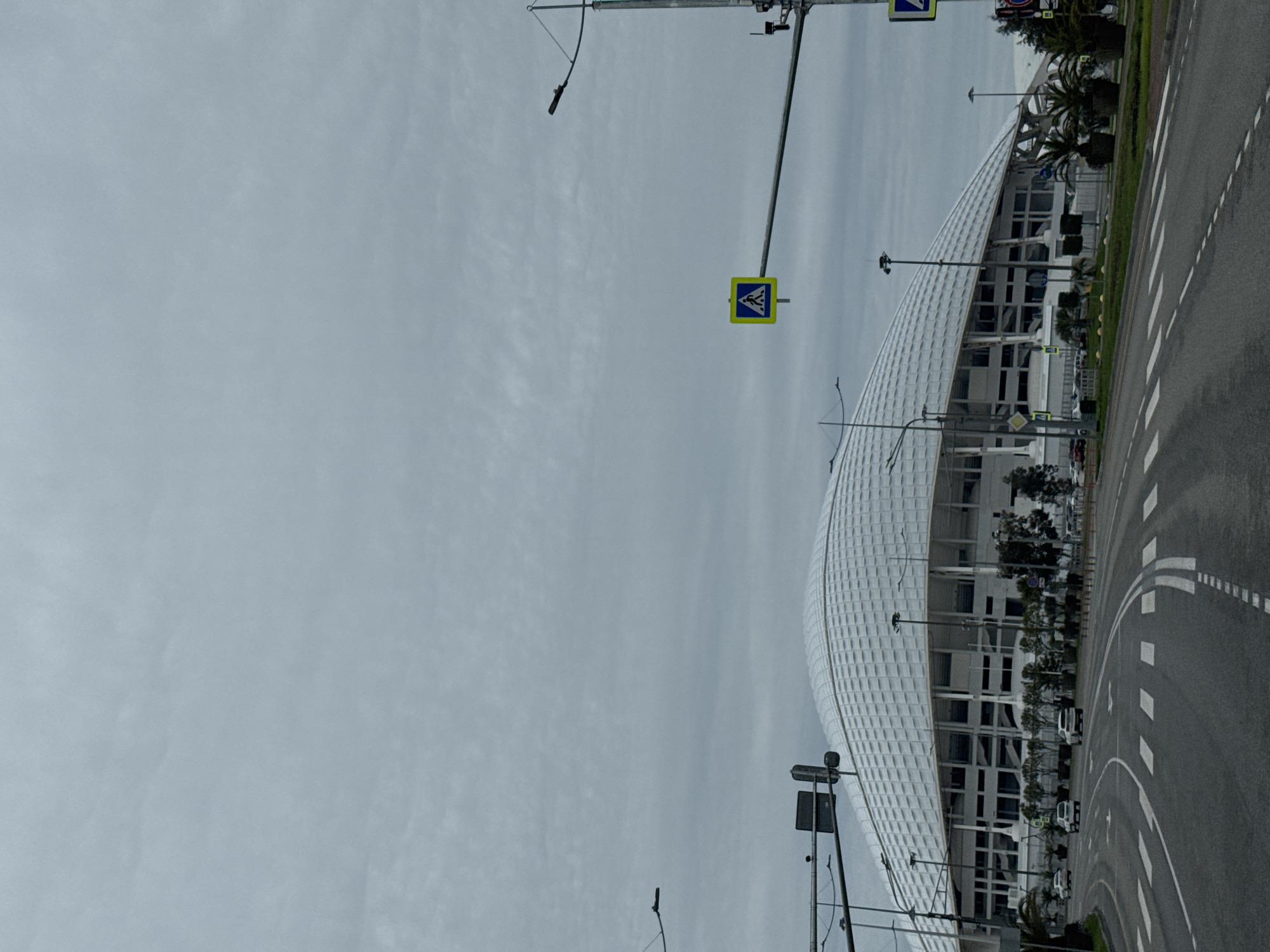 The image presents a low-angle, wide-angle view of Fisht Olympic Stadium in Sirius, Russia, on an overcast day. The stadium, characterized by its white, patterned outer shell and arched structure, dominates the right side of the frame. Adjacent to the stadium is a paved road with painted white lane markings and a green strip of vegetation. A pedestrian crossing sign is visible atop a traffic pole in the foreground. Multiple light poles extend upwards against the backdrop of the gray, cloudy sky, which constitutes the majority of the image's left half.