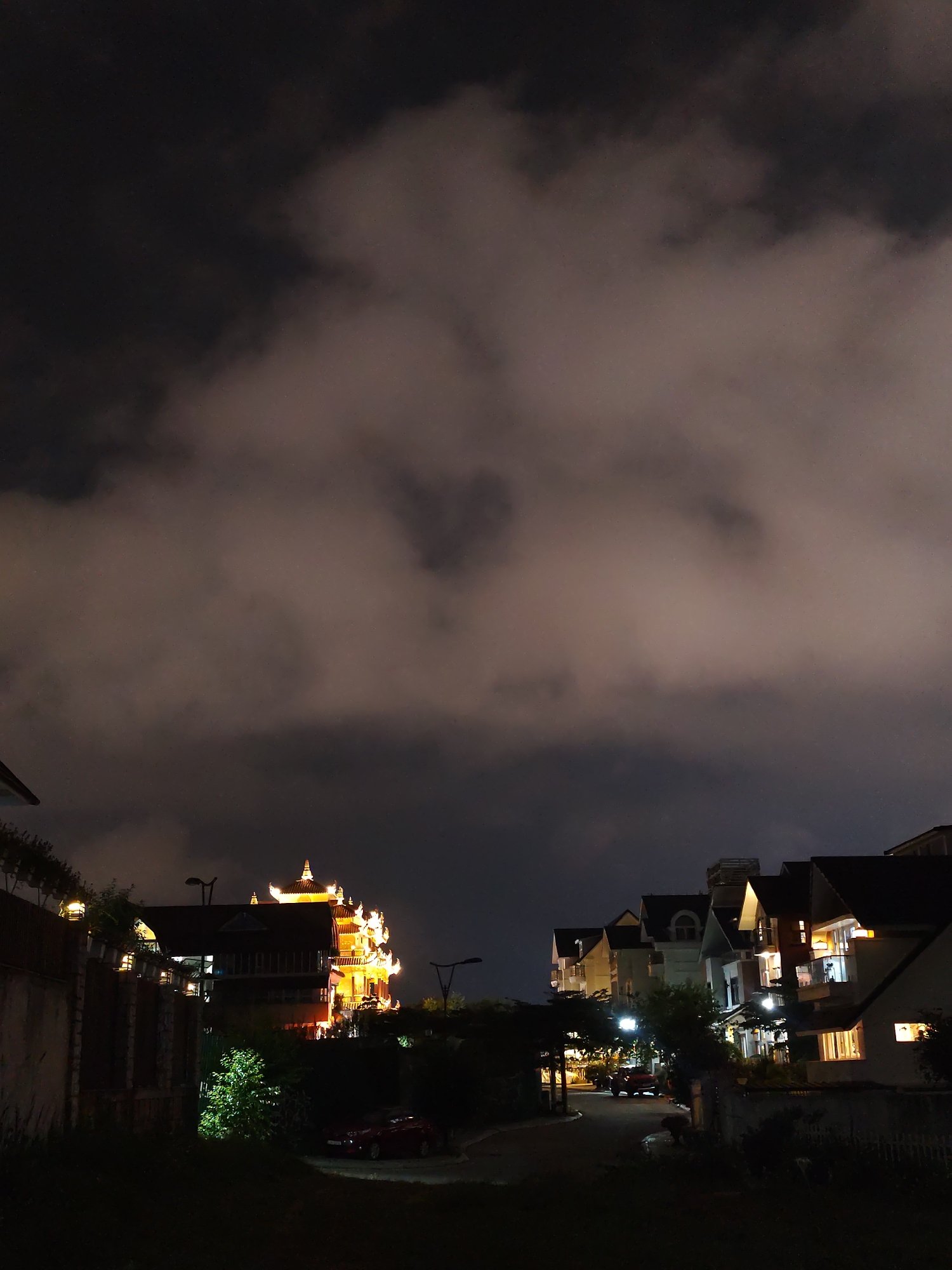 The image depicts a night view of a residential area in Da Lat, Vietnam. The foreground shows a street winding between houses with dark roofs. Streetlights illuminate the scene. On the left, a wall runs alongside a multi-story building with ornate, brightly lit architectural details resembling a pagoda. A car is parked near the building. On the right, a row of houses with varying architectural styles is partially visible, some with illuminated windows. The sky is filled with heavy, dark clouds.
