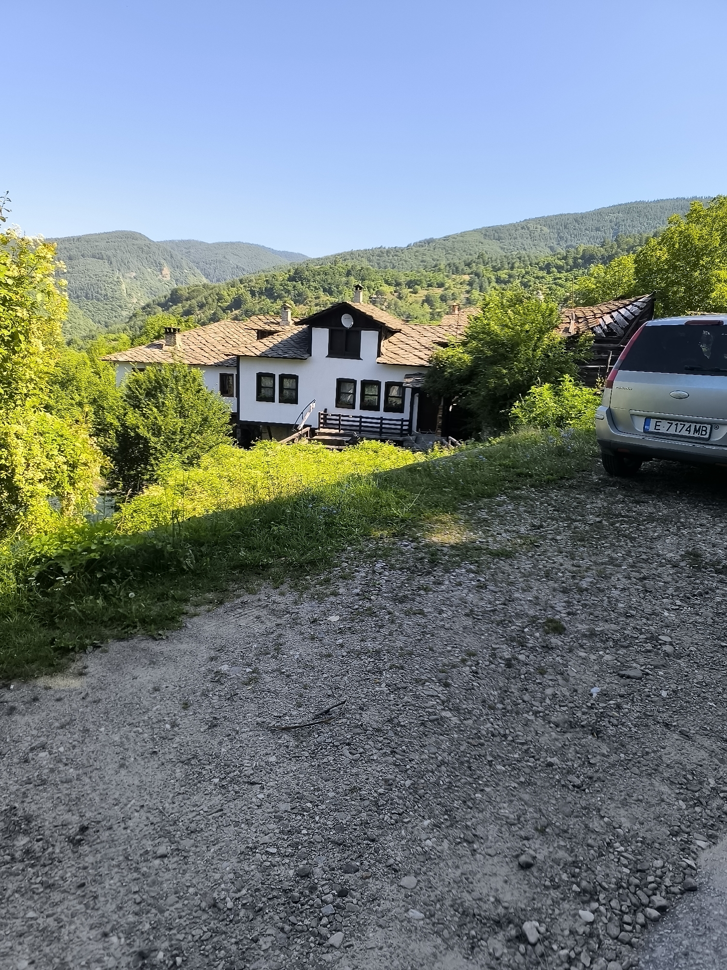 A silver Ford Fusion, license plate E 7174 MB, is parked on a gravel driveway in front of a traditional white house with a stone-tiled roof. The house is situated on a hillside surrounded by lush green vegetation and rolling, tree-covered mountains under a clear blue sky. The scene appears to be in Kovachevitsa, Bulgaria, a village known for its well-preserved Revival-era architecture. The image captures a serene rural setting bathed in warm sunlight.