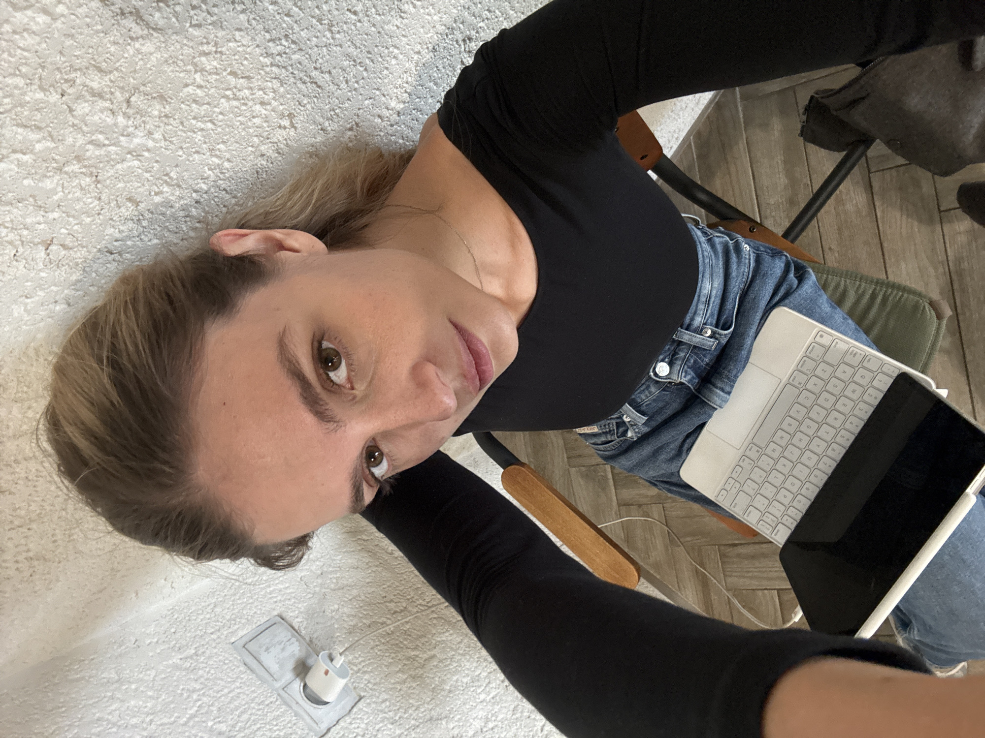 A high-angle portrait shows a woman sitting indoors, looking up towards the camera. The image is oriented sideways, giving the impression of her lying down. She has light blonde hair, light brown eyes, and fair skin, with a natural makeup look including pinkish lipstick. She is wearing a fitted black long-sleeved top and blue denim jeans with a "DIESEL" logo visible on the back pocket.
She holds a silver-colored tablet or laptop with a white keyboard attachment, likely an iPad with a Magic Keyboard, resting on her lap. The screen of the device is dark. She is seated in a chair with a green fabric seat and wooden and dark metal frame elements.
The background features a textured, off-white wall on her left and a light-colored wooden plank floor on her right. A white power adapter is plugged into a wall outlet near the bottom left of the frame. Soft, even lighting illuminates the scene. The overall atmosphere is casual and relaxed.