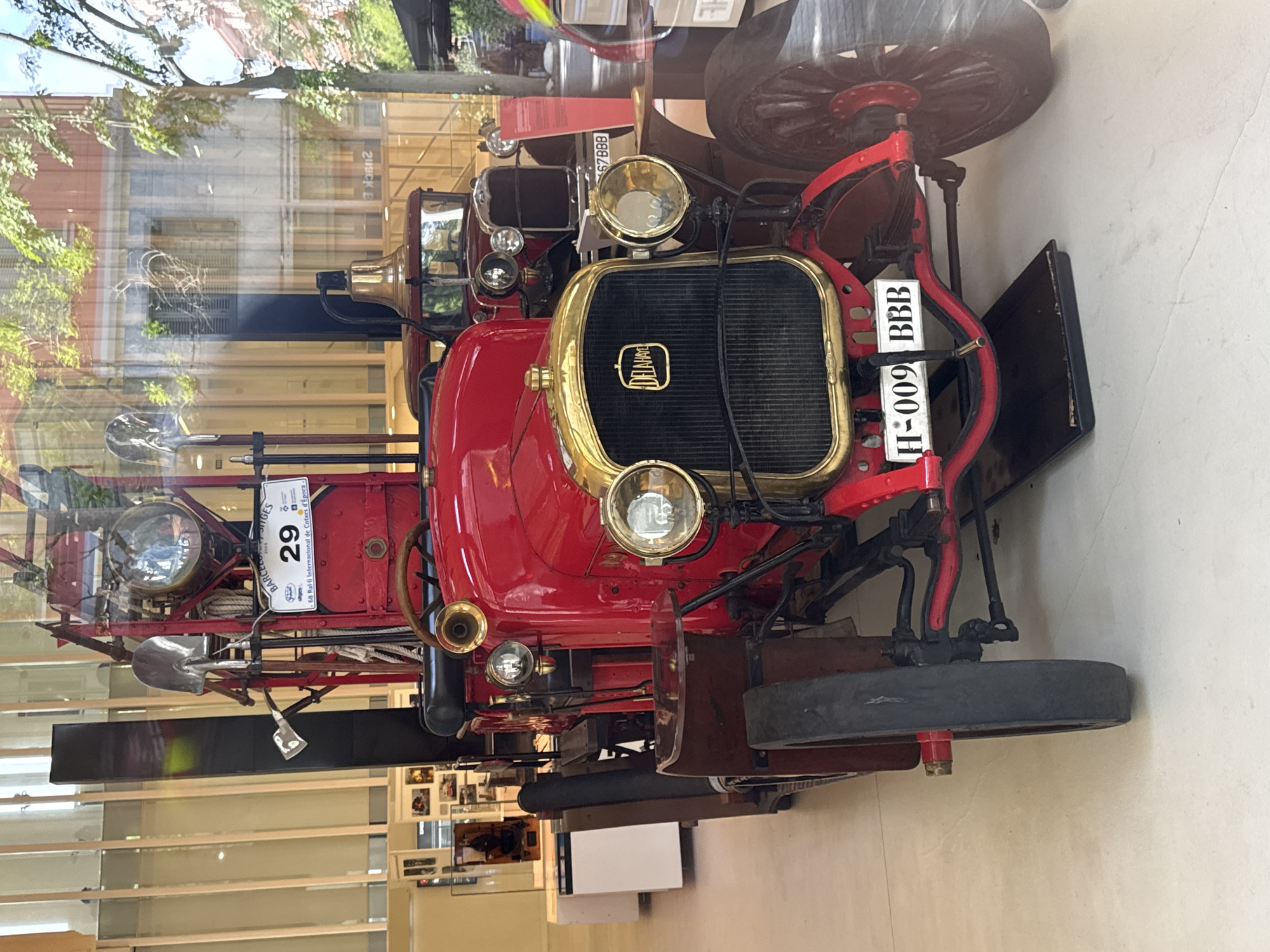 A vintage, bright red vehicle, likely a fire engine or utility car, is the central subject of this image. It is displayed indoors, possibly in a museum or exhibition hall, with reflections of trees and buildings visible on a large window or glass enclosure in the background. The vehicle features prominent brass-colored accents, including a large grille with the "DELAHAYE" logo, multiple headlights, and a horn. Its large wheels have wooden spokes.
Tools like a shovel and an axe are mounted on the side of the vehicle, suggesting its utilitarian purpose. A white event tag with the number "29" is attached, along with text that reads "BARCELONA SITGES" and "68 Ral-li Internacional de Cotxes d'Época", indicating its participation in the 68th International Vintage Car Rally, likely in Barcelona, Spain. The date "2026" is also partially visible on this tag. The front license plate clearly displays "H-009 BBB". Another partially visible license plate in the background reads "67BBB", and reflected text shows "209CK B". The vehicle appears meticulously maintained and polished, consistent with an exhibit. The bright lighting suggests it is daytime.