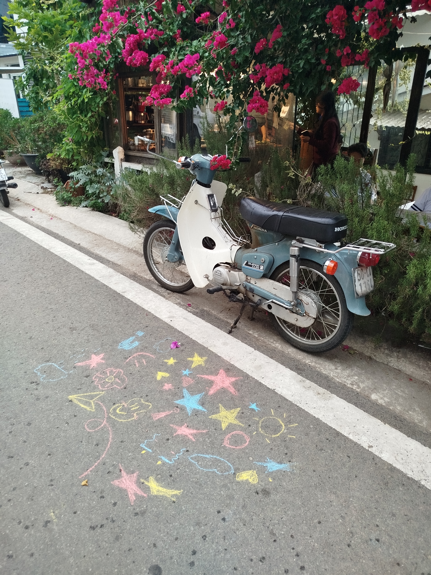 The scene is in Da Lat, Vietnam, featuring a vintage motorcycle parked beside a sidewalk. The motorcycle is light blue and white, bearing the brand "Honda." A chrome luggage rack is attached behind the seat. The license plate, partially obscured, reads "50-CA." On the asphalt street, brightly colored chalk drawings of stars, a sun, flowers, clouds, and other whimsical shapes are visible. The background shows a building adorned with vibrant pink bougainvillea flowers. A person in dark clothing is standing near a window, while others are seated nearby. Lush greenery and potted plants surround the building. The sidewalk is defined by a white line marking the edge of the road.