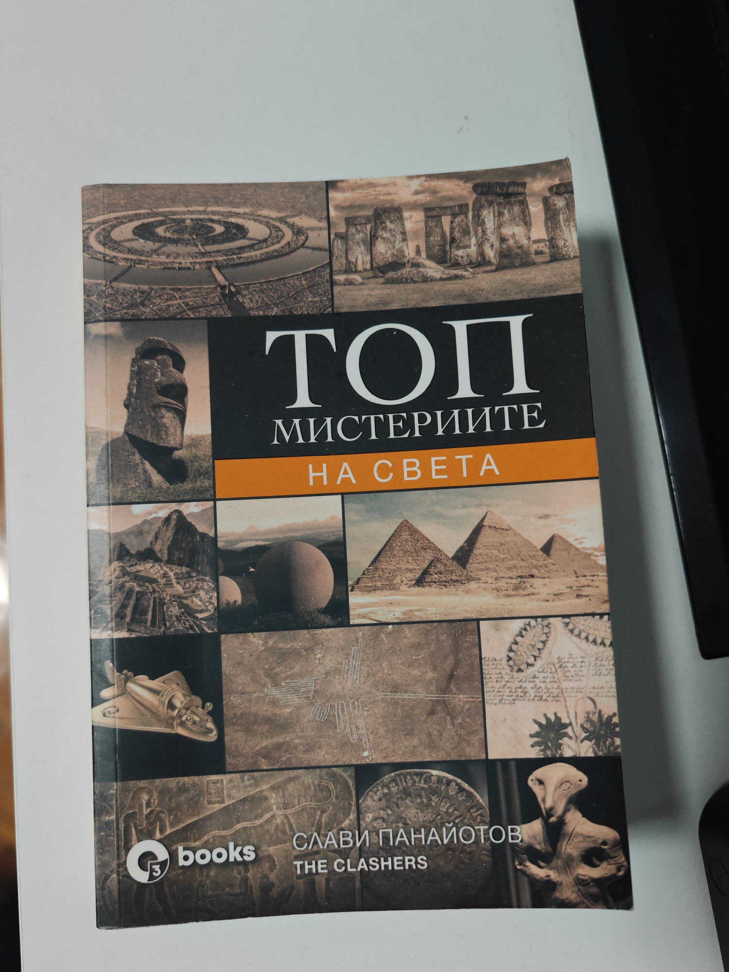 A book titled "ТОП МИСТЕРИИТЕ НА СВЕТА" (TOP MYSTERIES OF THE WORLD) by Слави Панайотов (Slavi Panayotov) and "THE CLASHERS," published by G3 books, is positioned diagonally on a clean, uniformly white surface. The book's cover features a multi-panel collage of nine distinct images associated with ancient mysteries: an aerial view of a circular settlement, Stonehenge, a Moai statue, the Giza pyramids, Machu Picchu, two large spherical stones, a golden winged artifact resembling an airplane, Nazca Lines, ancient textual diagrams, and an Ubaid lizard-like figurine. An additional ancient relief carving is partially visible at the bottom left behind the publisher's logo. A dark, vertical rectangular object, likely a component of an electronic device, is partially visible along the right edge of the frame. The specific location within Shumen, Bulgaria, cannot be determined from the visual information provided.