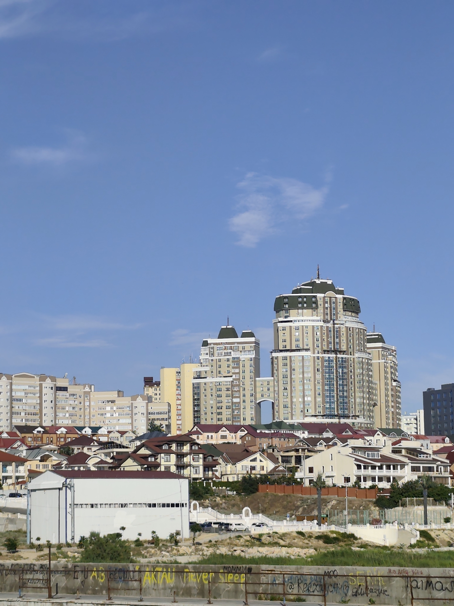 This media file captures a cityscape under a bright, clear blue sky with scattered wisps of clouds, suggesting a sunny day. The scene is dominated by a variety of buildings, ranging from low-rise houses with red-tiled roofs to mid-rise apartment blocks, and prominently featuring several tall, modern high-rise buildings in the background. The architecture of the high-rises is characterized by a palette of light brown, beige, and white, with dark windows and decorative accents. One particularly distinctive tower has a multi-tiered, green-roofed crown.
In the foreground, a wide embankment is visible, constructed from concrete and featuring a metal railing. The embankment's surface is covered in graffiti. Some of the visible text, in yellow and white spray paint, reads "AKTAU Never Sleep", "mommia", and other illegible markings. This text, along with the general architectural style, hints at a location in Aktau, Kazakhstan. The overall setting appears to be a coastal or waterfront urban area, with the embankment suggesting a proximity to water. No people are clearly discernible in the image, and no specific activity or event is taking place. The scene is static, focusing on the urban landscape.