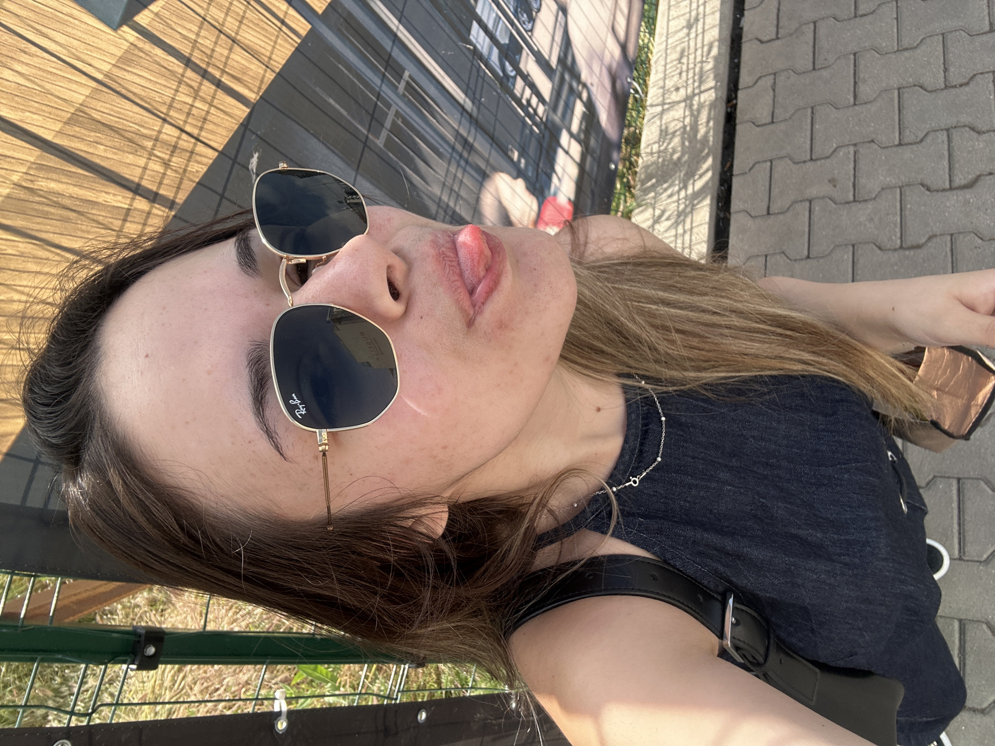A close-up, high-angle selfie captures a person outdoors on a sunny day in Sofia, Bulgaria. The image is rotated on its side, focusing on the subject's face and upper body. The person, with long, light brown hair, fair skin, and freckles, is wearing dark, square-framed sunglasses with "Ray-Ban" visible on the left lens. Their tongue is playfully sticking out, conveying a cheeky or relaxed expression.
The individual is wearing a dark-colored, possibly textured, top and a thin silver necklace adorned with small beads and a key-shaped pendant. A black strap, likely from a bag, is visible over their shoulder.
The background elements suggest an outdoor, semi-urban setting with bright, direct sunlight creating strong shadows. To the upper left, a wooden or slatted surface with prominent linear shadows is visible. Behind the subject's head, there's a blurred building or large structure. On the right, gray interlocking paver bricks form a paved area. In the lower section, a green metal fence or railing with some grass behind it can be seen. The overall scene is bright and casual.