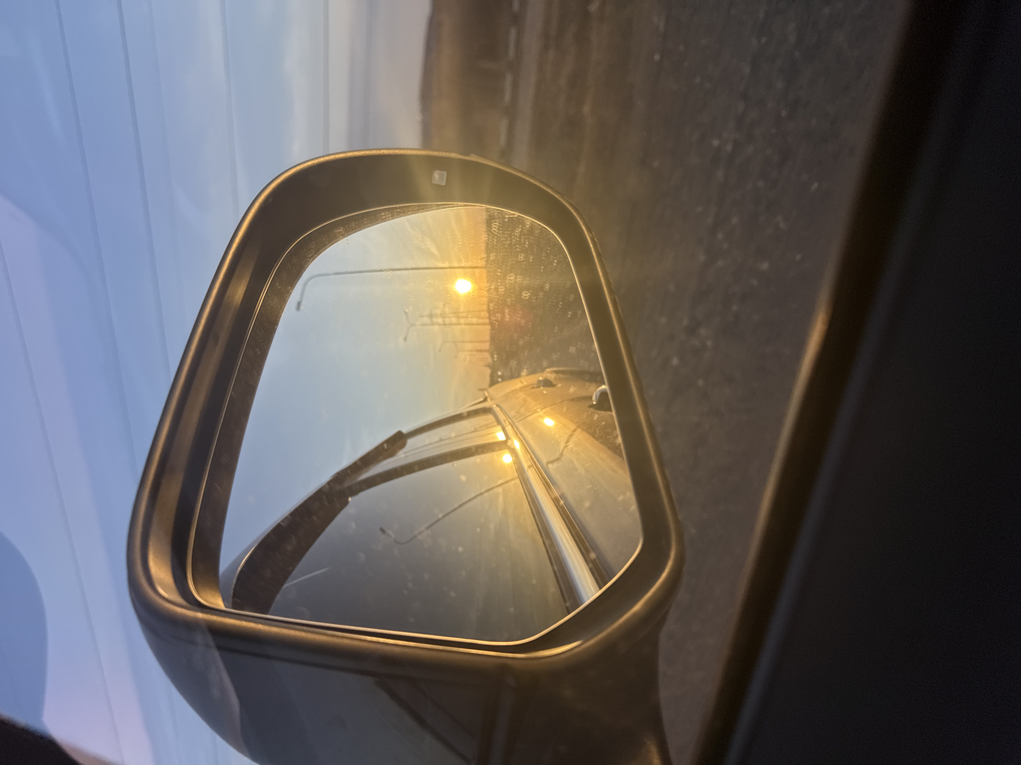 The image shows a car's side mirror reflecting a road illuminated by streetlights, likely at dusk or dawn. The road markings are visible. The setting is assumed to be in or near Chaikovsky, Russia. There are no people visible. The focus is on the mirror and the road it reflects. The sky is visible in the background, fading from blue to light.