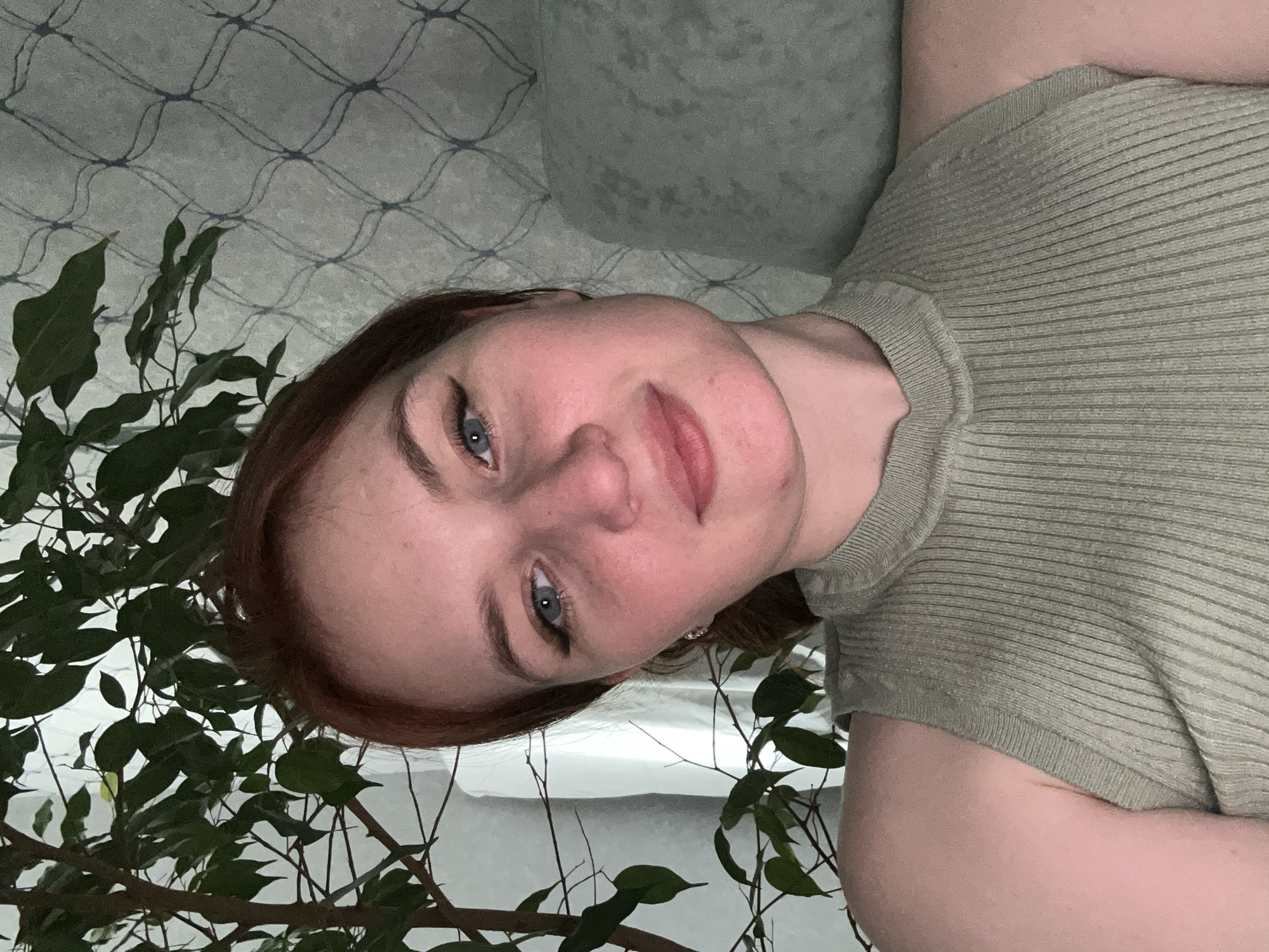 The image displays a close-up, chest-up portrait of a young woman, rotated 90 degrees counter-clockwise. She has fair skin, reddish-brown hair, and striking blue eyes, looking directly at the camera with a serene, slightly smiling expression. Her eyebrows are well-defined, and she has minimal visible makeup. She is wearing a short-sleeved, light olive green or khaki-colored top with a distinct ribbed texture.
The setting is indoors, likely a home environment in Chaikovsky, Russia. A vibrant green houseplant with numerous leaves and thin branches is prominent on the left side and bottom of the frame, partially framing the woman. In the background, a light-colored, possibly grey, wall or wallpaper features a subtle, repeating geometric pattern resembling a crisscrossing lattice or net. A soft, light-colored pillow or cushion is visible behind her head on the right side of the frame. The lighting is soft and diffused, creating a gentle and even illumination without harsh shadows, consistent with an indoor setting. No text is visible in the image.