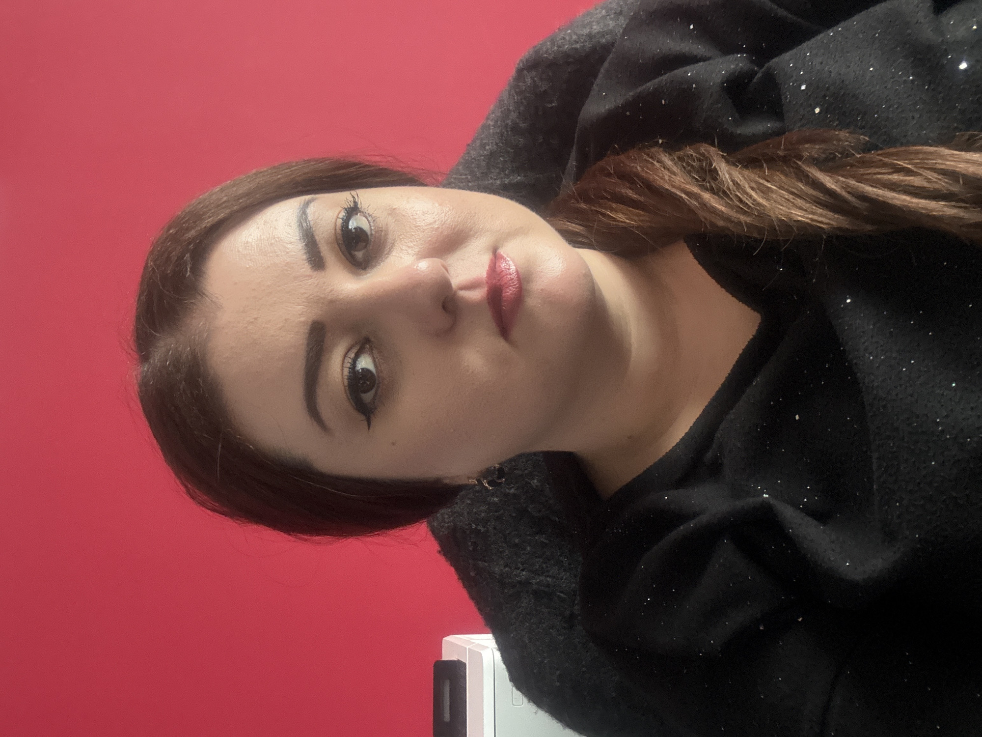 A woman is centered in a selfie with a red wall background. She has brown hair, fair skin, and is wearing a dark-colored, sparkly sweater. Her hair is styled to the side, and she is wearing makeup, including eyeliner and lipstick. The bottom of a white object is partially visible in the lower-left corner. There is no indication of the location being Varna, Bulgaria.
