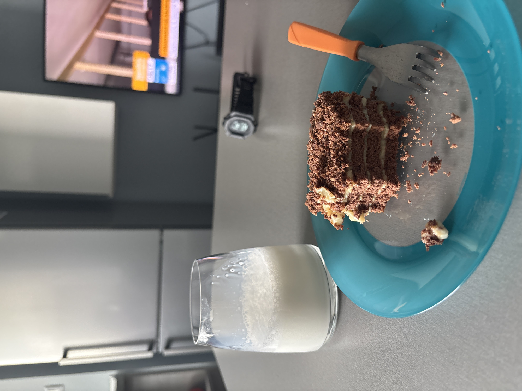 A partially eaten slice of chocolate cake sits on a blue plate with an orange-handled fork. A glass of milk is beside the plate. In the background is a television and a grey wall with a mounted object. There is white kitchen cabinetry. The city and country are not discernible from the image.