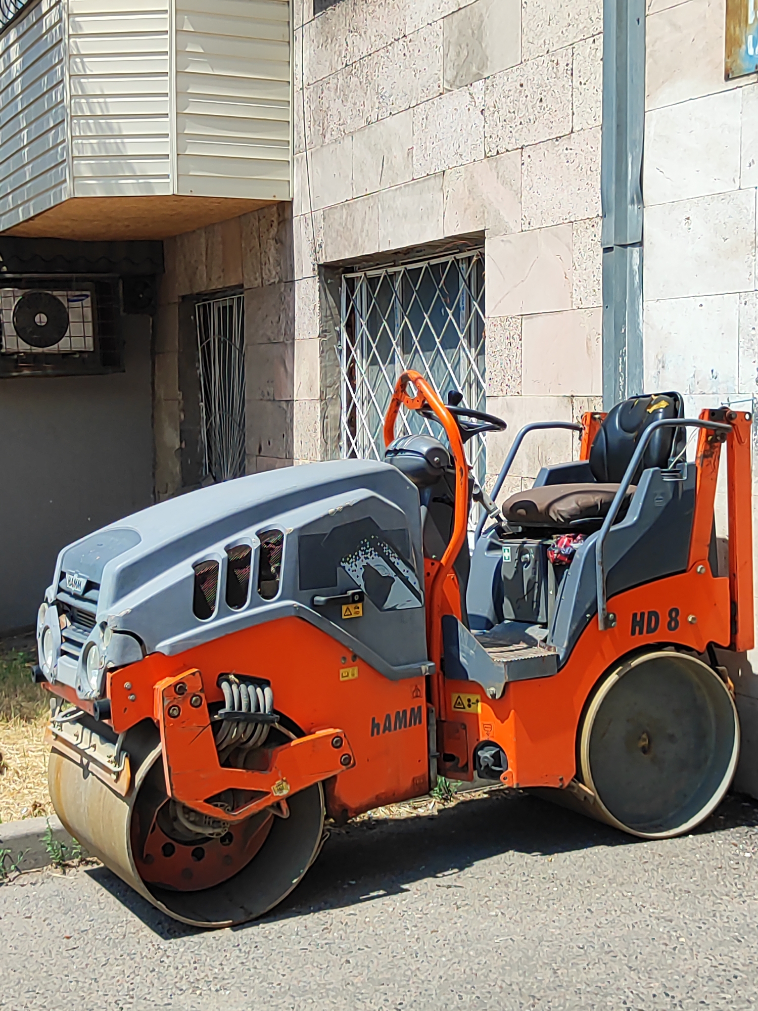 This image shows an orange and gray Hamm HD 8 road roller parked on an asphalt surface next to a building. The building has a light-colored stone facade and a white, horizontal slatted overhang. There is an air conditioning unit mounted on the building wall to the left of the road roller. The road roller has a prominent front drum and a rear drum, with a seat and steering wheel for an operator. The brand name "Hamm" is visible on the front of the machine, and "HD 8" is visible on the rear. The scene appears to be outdoors during the daytime, with sunlight casting shadows. The environment suggests an urban or construction setting in Almaty, Kazakhstan. There are no people visible in the image, and the road roller is stationary, implying it is either parked or not currently in use. The overall mood is neutral, focusing on the industrial equipment and its surroundings.