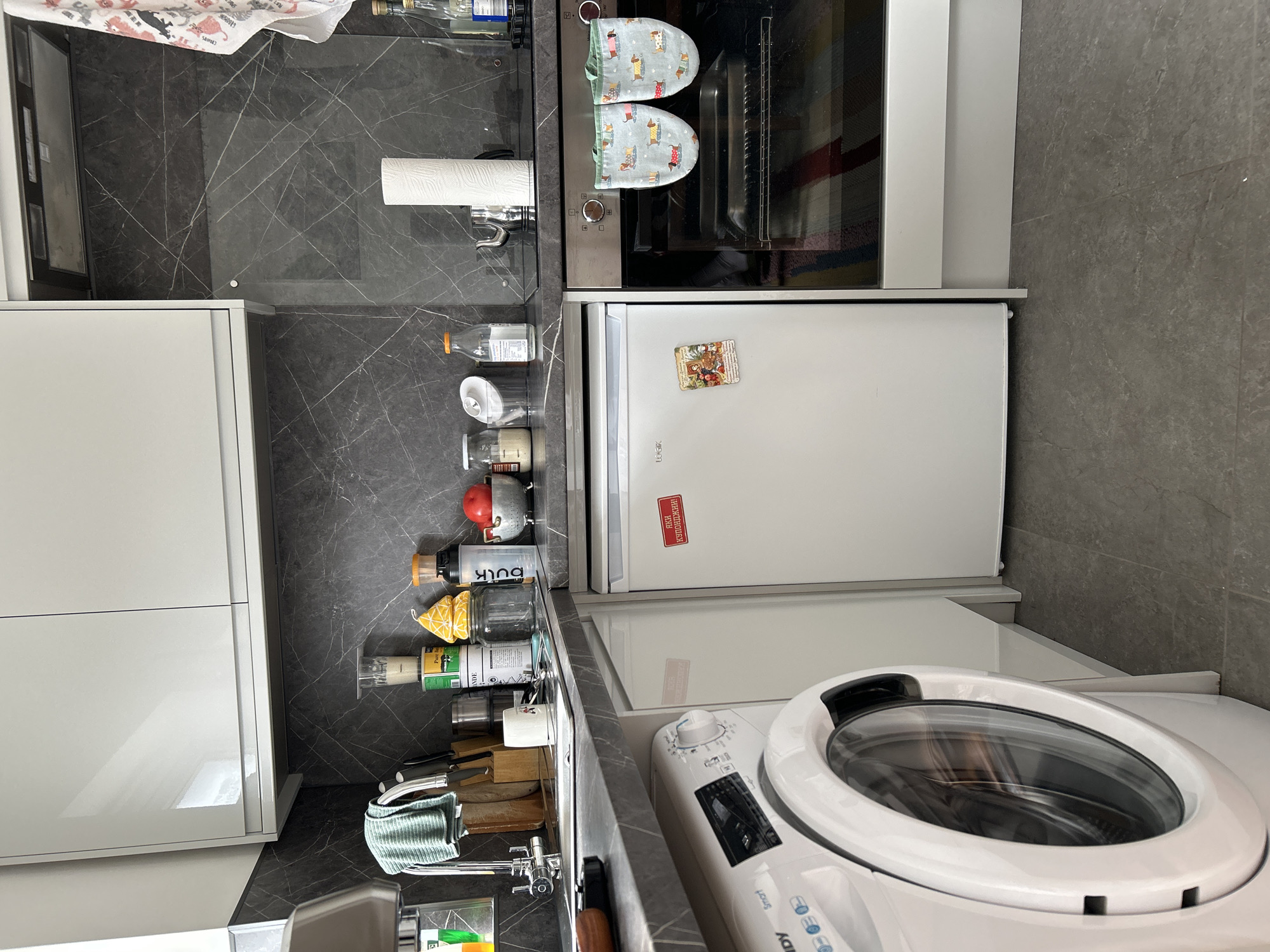 A close-up shot inside a kitchen shows a washing machine, a refrigerator, an oven, a countertop with various items, and cabinets. The washing machine is in the lower right corner, while the refrigerator is directly above it. Above the refrigerator is an oven with oven mitts on top. To the left of the oven, a countertop with jars, bottles, a paper towel dispenser, a knife block, and kitchen utensils is visible. To the left of the countertop are white cabinets. The backsplash and flooring are a gray-tone marble-like material. The location is a domestic kitchen, likely in the city of London, United Kingdom. The image contains no people or interactions.