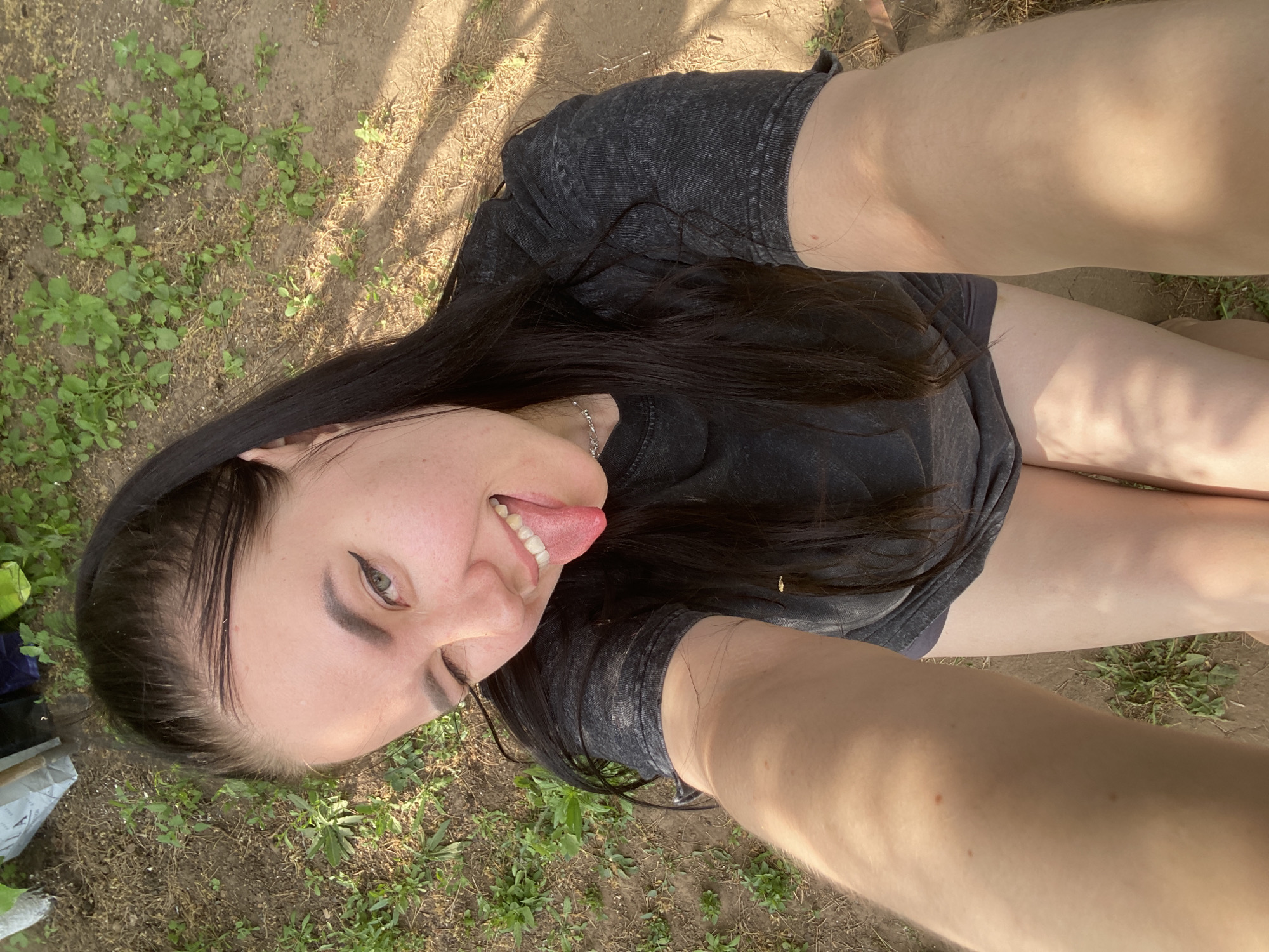 A woman is lying down in an outdoor location with grass and soil ground cover. She is wearing a dark-colored T-shirt and dark shorts. Her long, dark hair is spread around her head. She is sticking her tongue out. The location is similar to a yard with grass and soil but there are no identifying features that indicate the specific city of Chaikovsky, Russia.