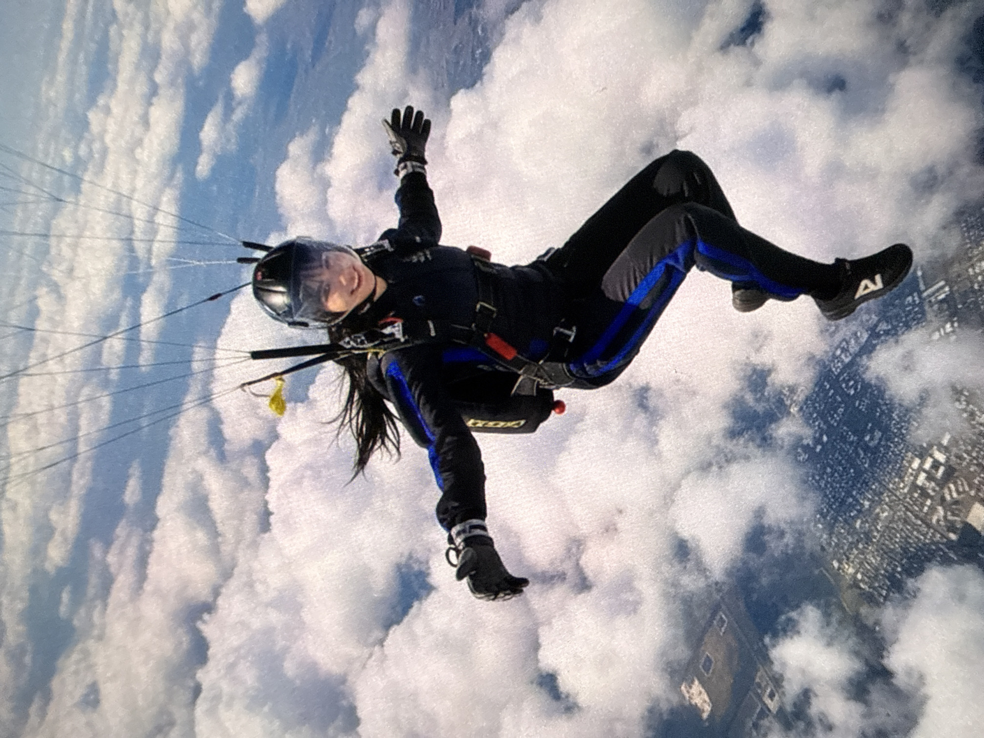 A woman is skydiving above a city. She is wearing a black jumpsuit with blue accents, a helmet, and gloves. The parachute strings are visible above her. The background consists of clouds and a cityscape.