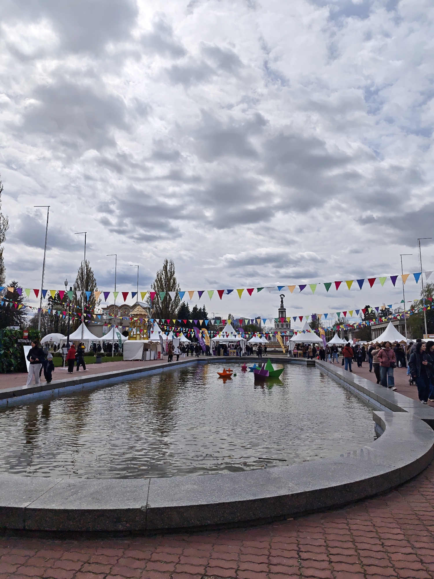 An outdoor market or festival is depicted on an overcast day in Kyiv, Ukraine. The scene is dominated by a long, curving fountain or pool in the foreground, filled with calm water that reflects the cloudy sky. Several large, colorful origami-style boat sculptures in orange, purple, and green float on the water.
Red-brown interlocking paving stones create pathways around the fountain, where numerous people are seen walking. White market tents and stalls line both sides of the fountain, indicating various vendors or exhibits. Above the bustling scene, strings of vibrant triangular pennant banners in red, yellow, green, blue, and purple stretch across the entire area, adding a festive atmosphere.
In the background, bare-branched trees suggest autumn or early spring. Utility poles rise above the tents. A prominent dark-colored statue on a light pedestal is visible in the mid-distance, nestled among the tents. Behind the tents, buildings with architectural details, including one with a decorative roof, can be seen. On the far left, a dark green sign with white text appears to read "BOTANICALS," possibly identifying a specific stall or area. The crowd is casually dressed in jackets and coats, suggesting cool temperatures under the cloudy sky.