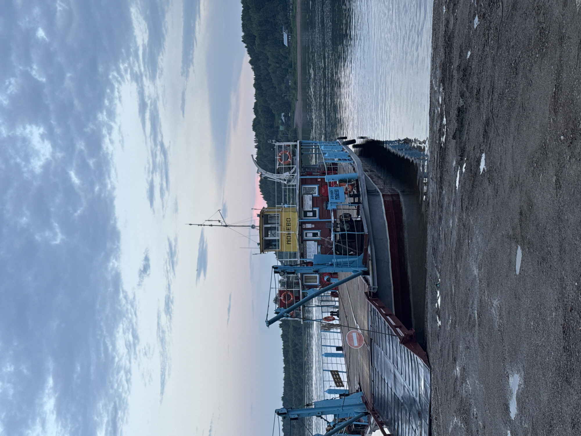 A ferryboat is docked at a muddy riverbank in Tyulkino, Russia. The boat is red, gray, and blue with a yellow cabin. The river is calm and reflects the overcast sky. The sky is a mix of gray and pink clouds. The riverbank is covered in mud and puddles. The scene appears to be at dawn or dusk.