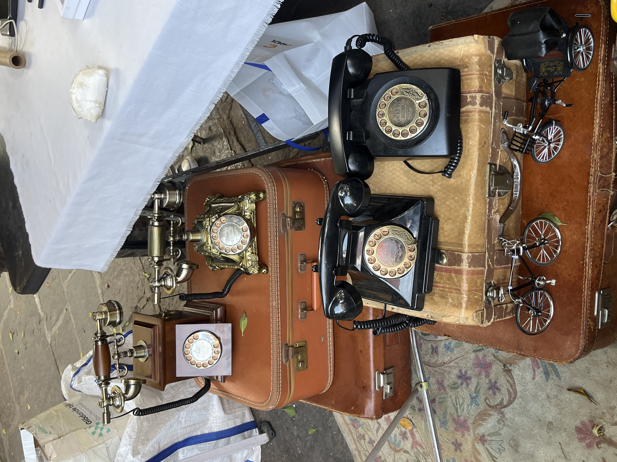 The image presents a collection of antique telephones and vintage suitcases displayed on what appears to be an outdoor sales table. Two rotary dial telephones, one black and one with brass accents, are prominently featured atop a stack of brown leather suitcases. A brass-colored telephone with a wooden base sits adjacent to the stack. Next to the telephones sits a miniature model of a bicycle on another stack of suitcases. The items are arranged against a backdrop that includes a white tablecloth and shopping bags, suggesting a market or flea market setting. The overall impression is that of a vintage or antique market. It is unclear whether this is in Puebla, Mexico.