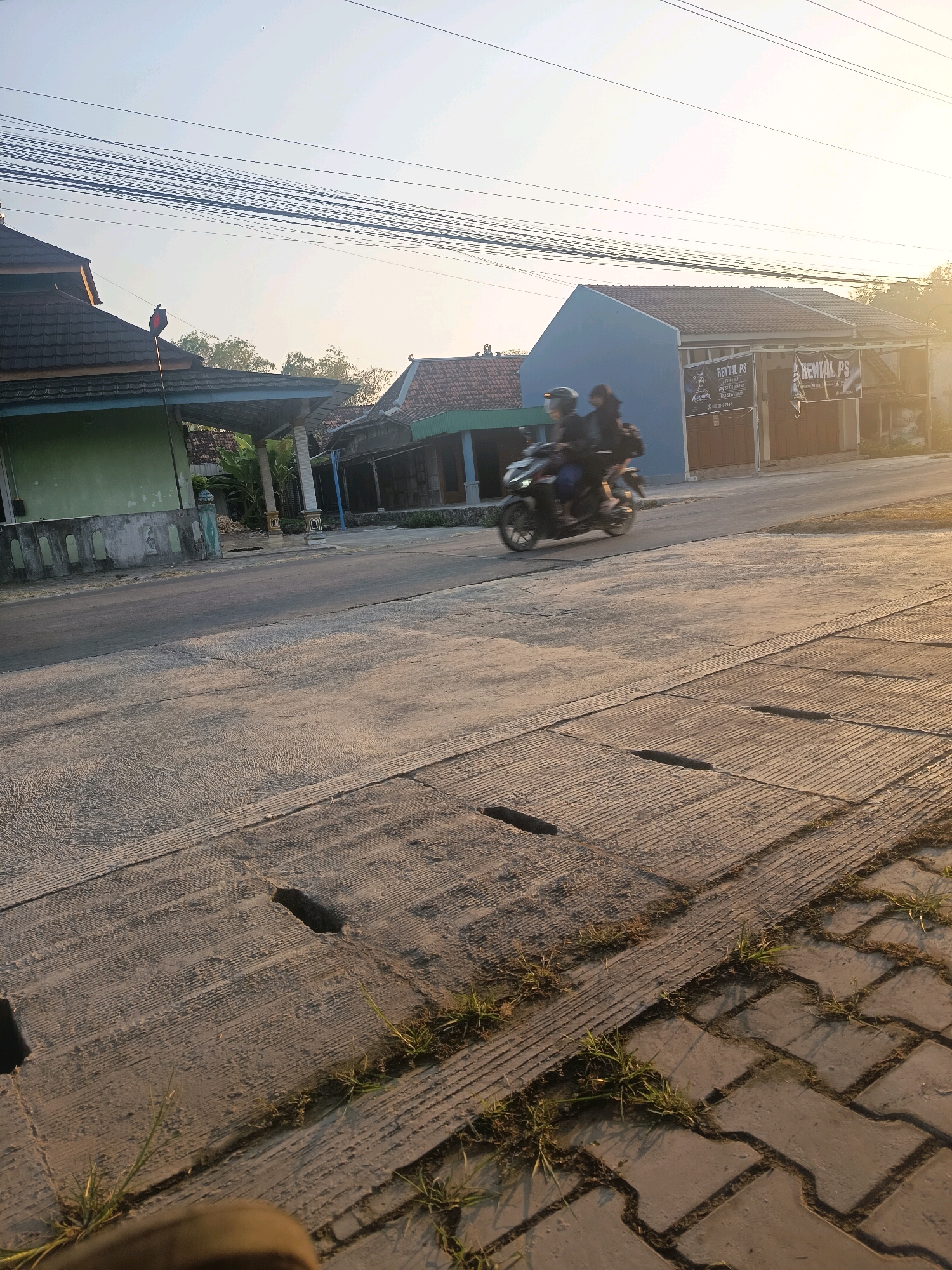 In Kalisari, Indonesia, the scene is illuminated by the golden light of a setting or rising sun, casting long shadows across a street. Two individuals, one driving and one as a passenger, are captured in motion on a scooter, their forms blurred by the speed. They are traveling on a paved road in front of a row of buildings. One building to the left is painted light green with a dark tiled roof and a red and black flag at its peak. To the right, a light blue building displays two banners for "RENTAL PS," indicating a PlayStation rental service. The foreground features a concrete sidewalk with interlocking pavers and a drainage grate. Vegetation, including grass, is growing between the pavers and along the edge of the road. Overhead, a complex network of electrical wires crisscrosses the sky, silhouetted against the bright sky. The environment suggests a residential or commercial street in a tropical locale.