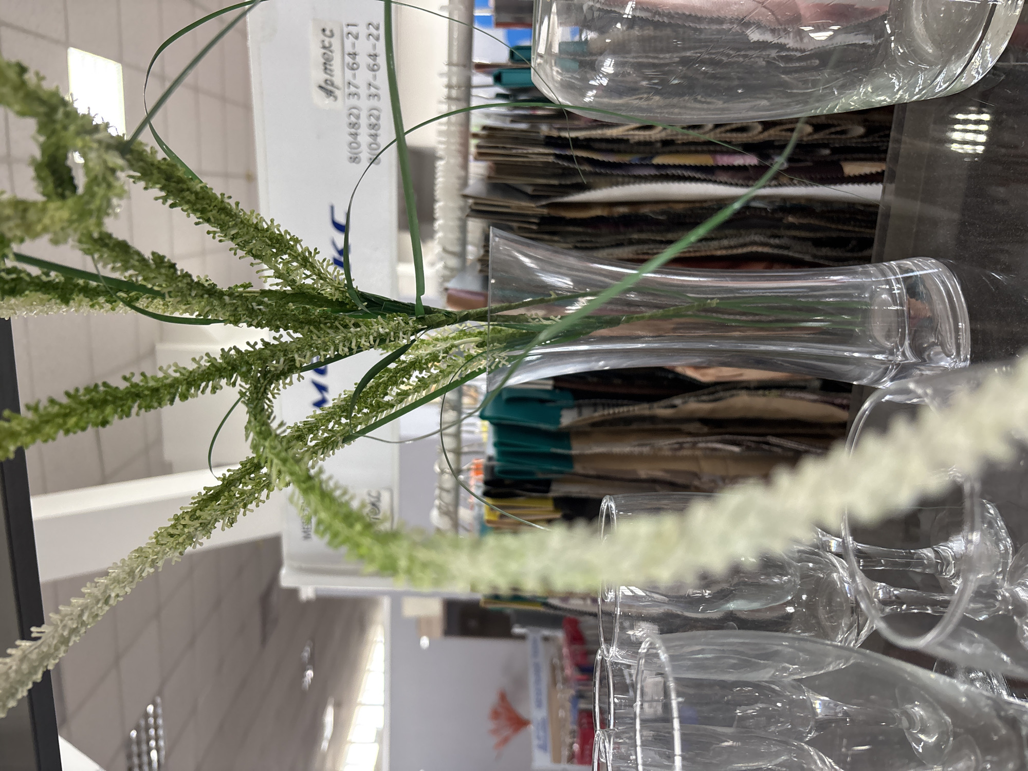 The image displays a close-up view of a clear glass vase containing a decorative artificial plant with green stalks and white feathery blooms. The vase is placed on a reflective dark surface, likely a countertop or table. In the background, a rack of fabric swatches is visible, suggesting a retail environment, possibly a fabric store or a showroom. Several other clear glass objects, possibly wine glasses or other stemware, are arranged around the vase in the foreground. A white sign with Russian text and a phone number is also visible in the background. The lighting appears to be artificial, casting reflections on the glass surfaces.
